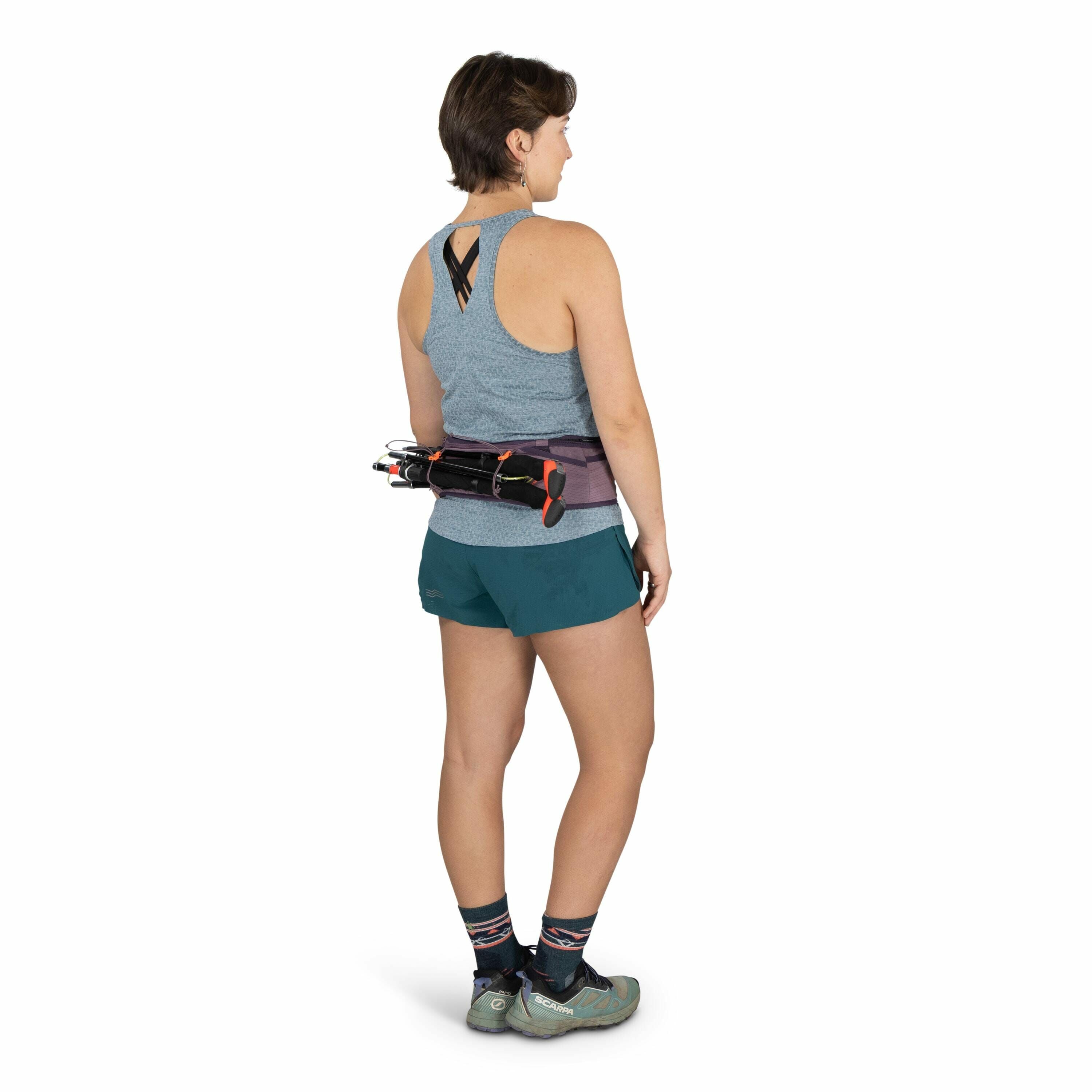The image depicts a person standing, facing away from the camera, wearing athletic gear suitable for outdoor activities. They are dressed in a light gray, sleeveless athletic top with a cross-strap design at the back and teal shorts. The individual is wea