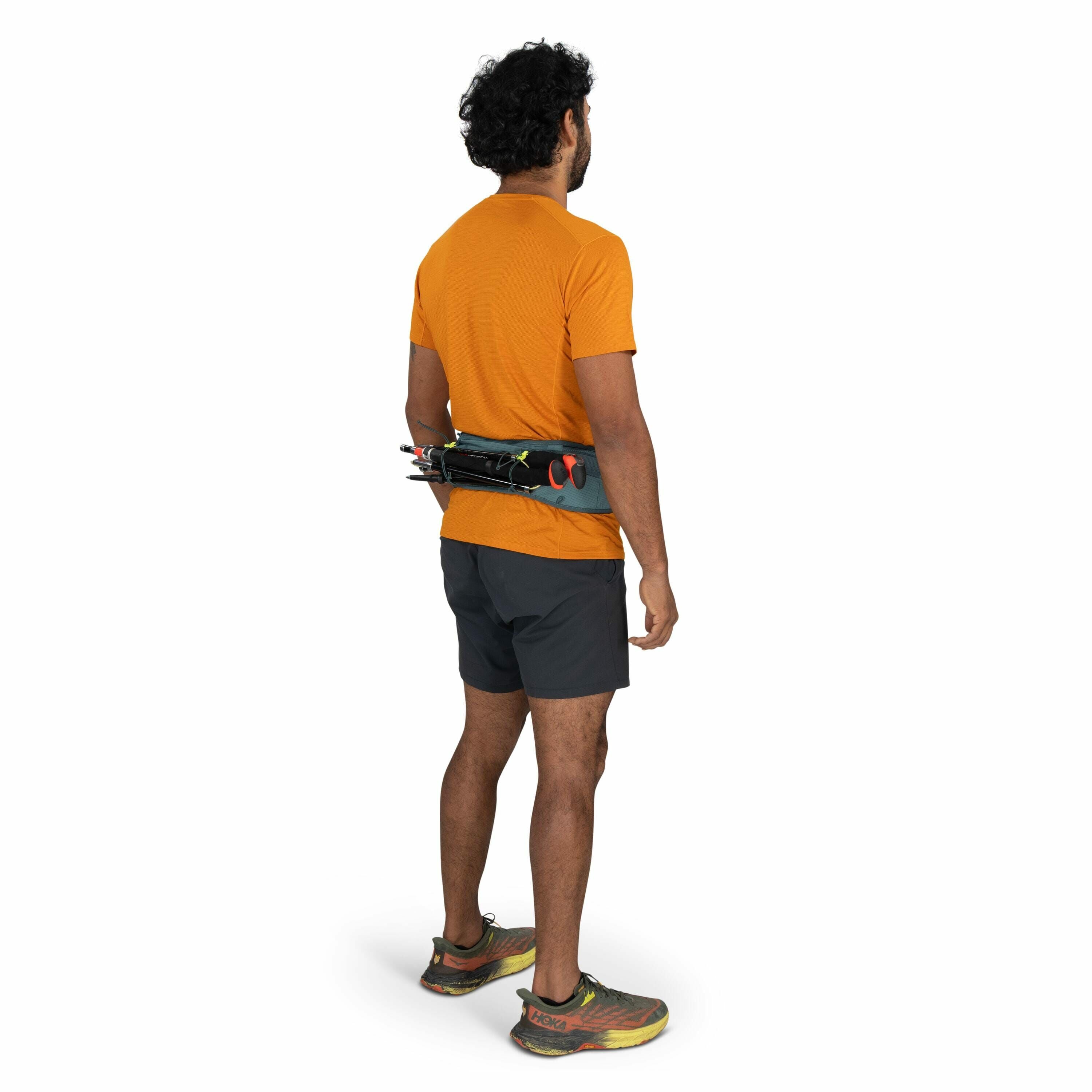 The image depicts a man standing and facing away from the camera, showcasing a runner's attire. He is wearing an orange short-sleeve shirt paired with dark gray shorts and multicolored running shoes that have visible Hoka branding. Around his waist, he ha