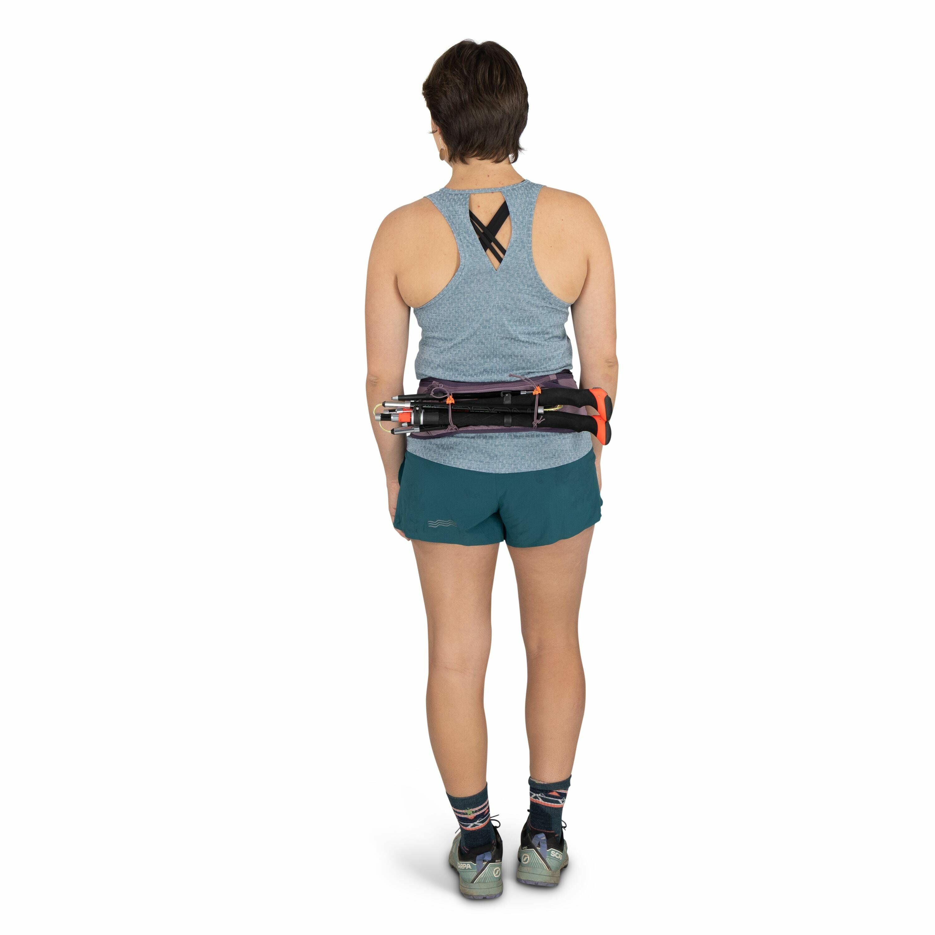 The image shows a person standing with their back to the camera. They are wearing a light blue sleeveless top with a black strap crisscrossing at the back, teal shorts, and gray athletic shoes. The person has a short hairstyle and is carrying a set of tre