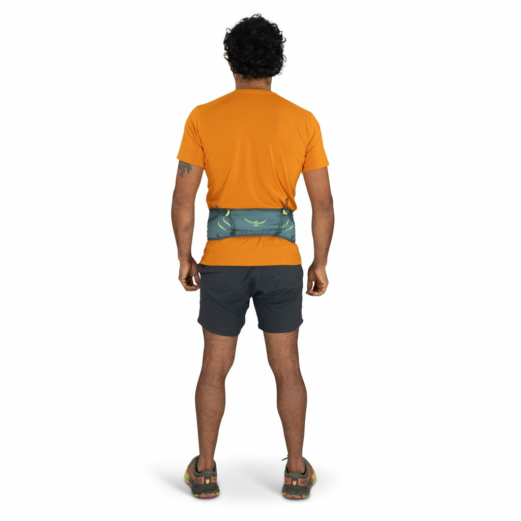 The images show a man standing with his back to the camera, wearing an orange T-shirt, black shorts, and trail shoes. Around his waist is a blue-green waist pack, which features a subtle pattern and a logo resembling a stylized bird with outstretched wing