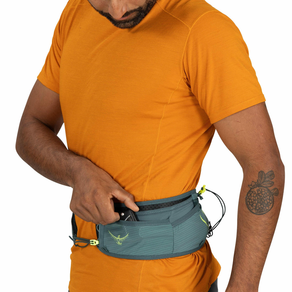 The image shows a man wearing an orange short-sleeved shirt and a gray waist pack with an Osprey logo on it. He is adjusting or reaching into one of the compartments of the waist pack. The waist pack has a green and yellow zipper with a toggle pull, provi