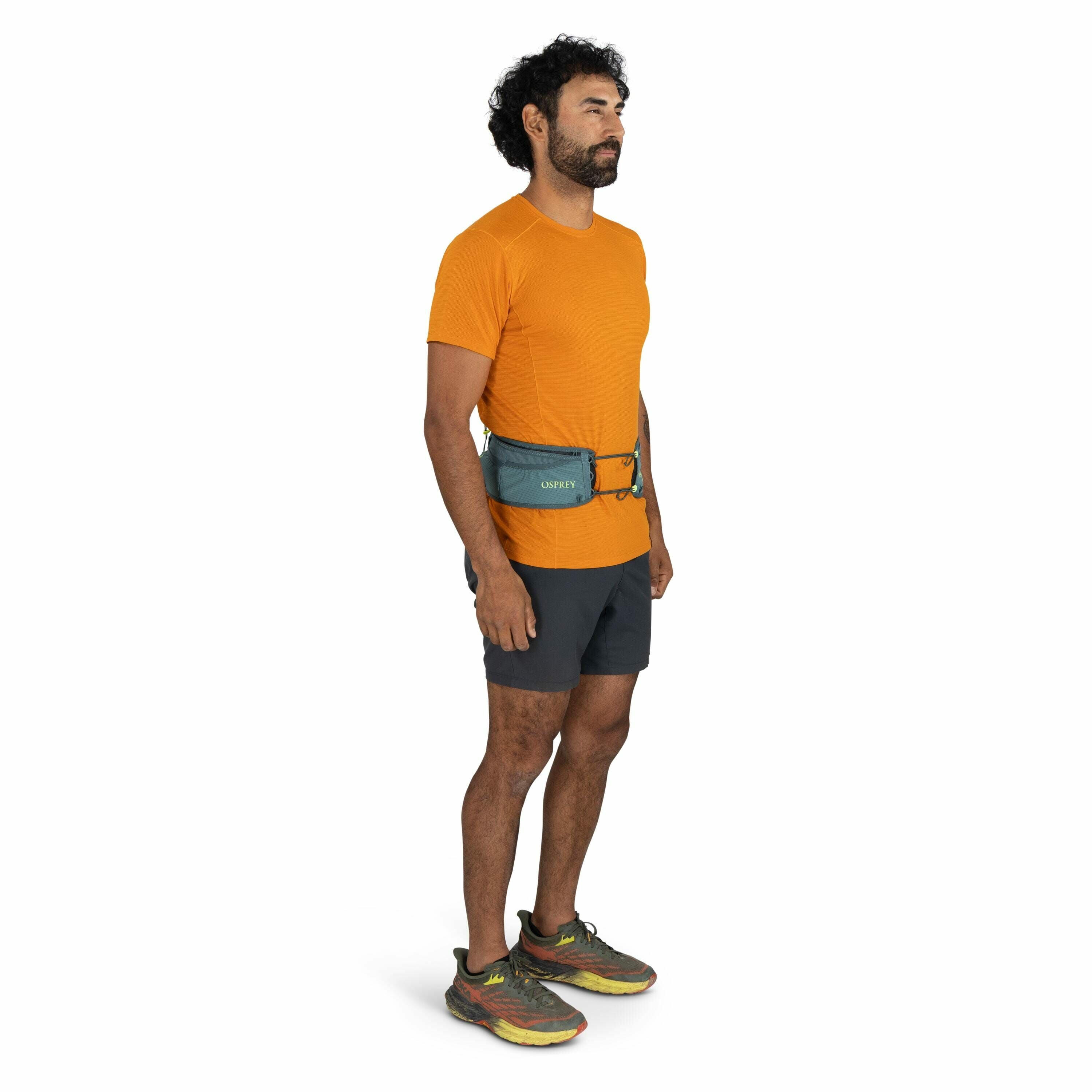 The image shows a person standing and dressed in sporty outdoor attire, ideal for activities like hiking or running. They are wearing an orange short-sleeve athletic shirt paired with dark gray shorts. Around their waist is a blue Osprey waist pack, which