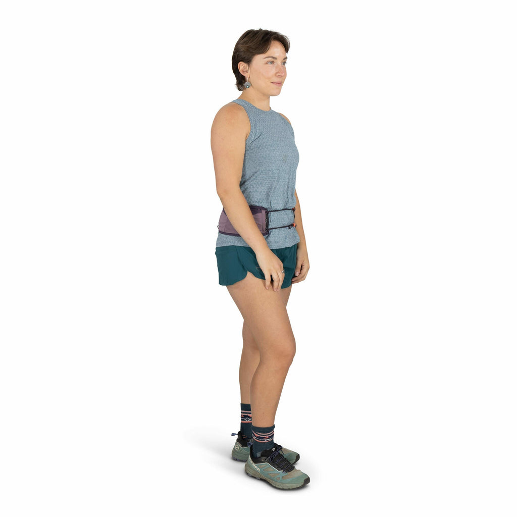 The image depicts a person standing in a side profile view, wearing a casual athletic outfit suitable for outdoor activities. They are dressed in a sleeveless, light blue textured top paired with teal athletic shorts. The person is wearing light blue and