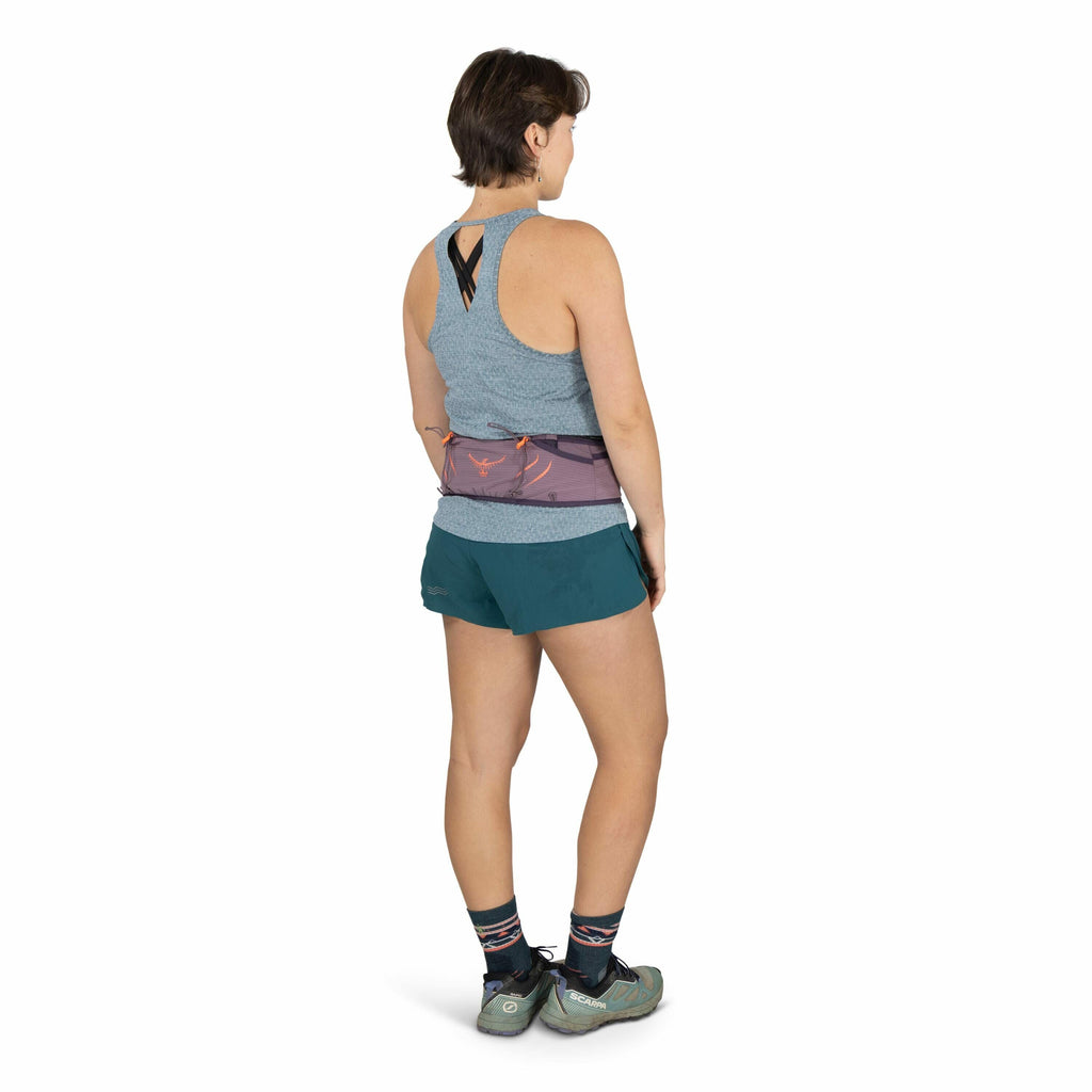 The image shows a woman standing with her back to the camera, wearing a casual athletic outfit. She is dressed in a light blue, textured, sleeveless top with black crisscross straps at the back, paired with teal shorts. The woman is wearing a purple waist