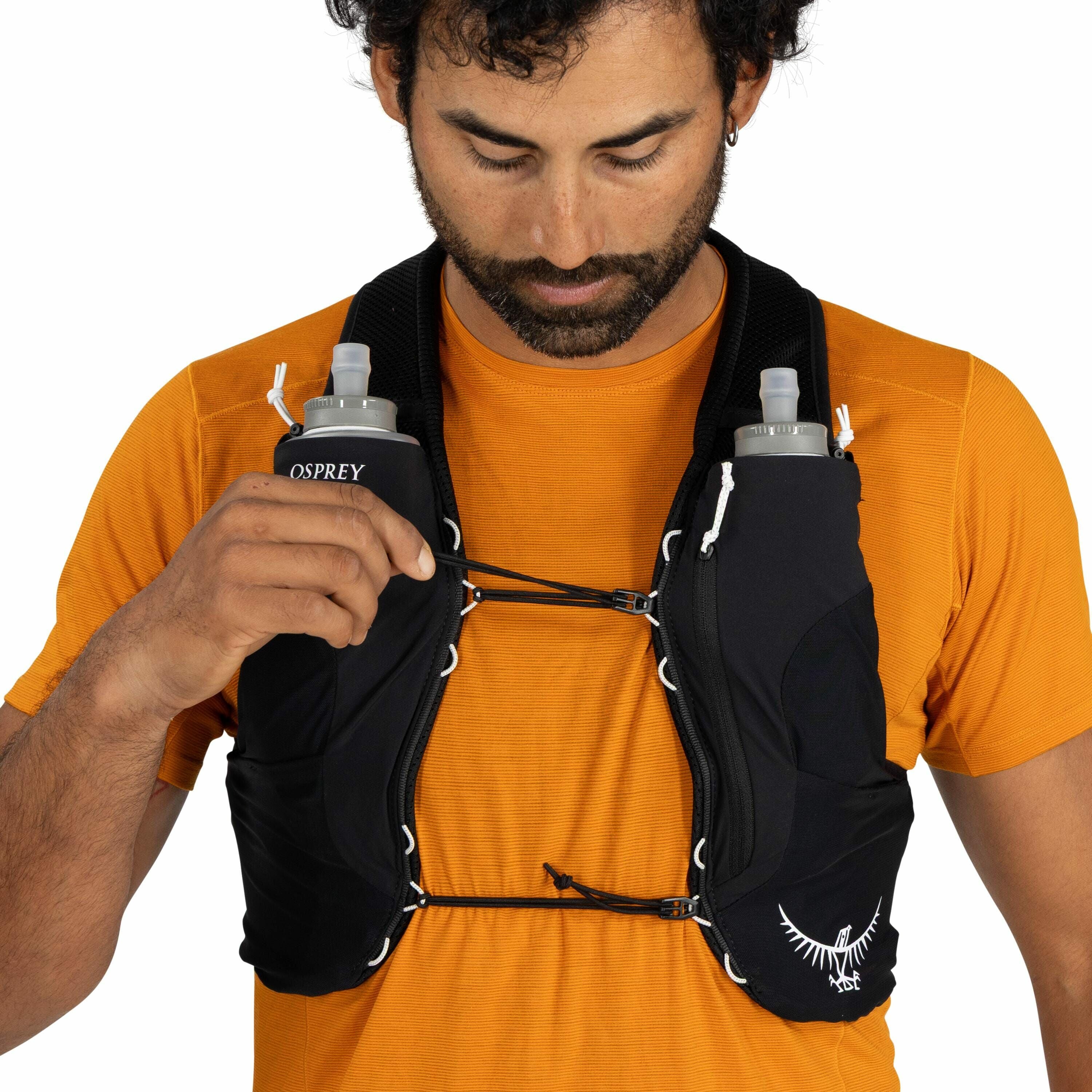 The image shows a person wearing a black hydration vest with two front pockets containing water bottles. The vest is secured with adjustable straps across the chest, allowing for a customizable fit. The person is wearing an orange shirt, providing a vibra