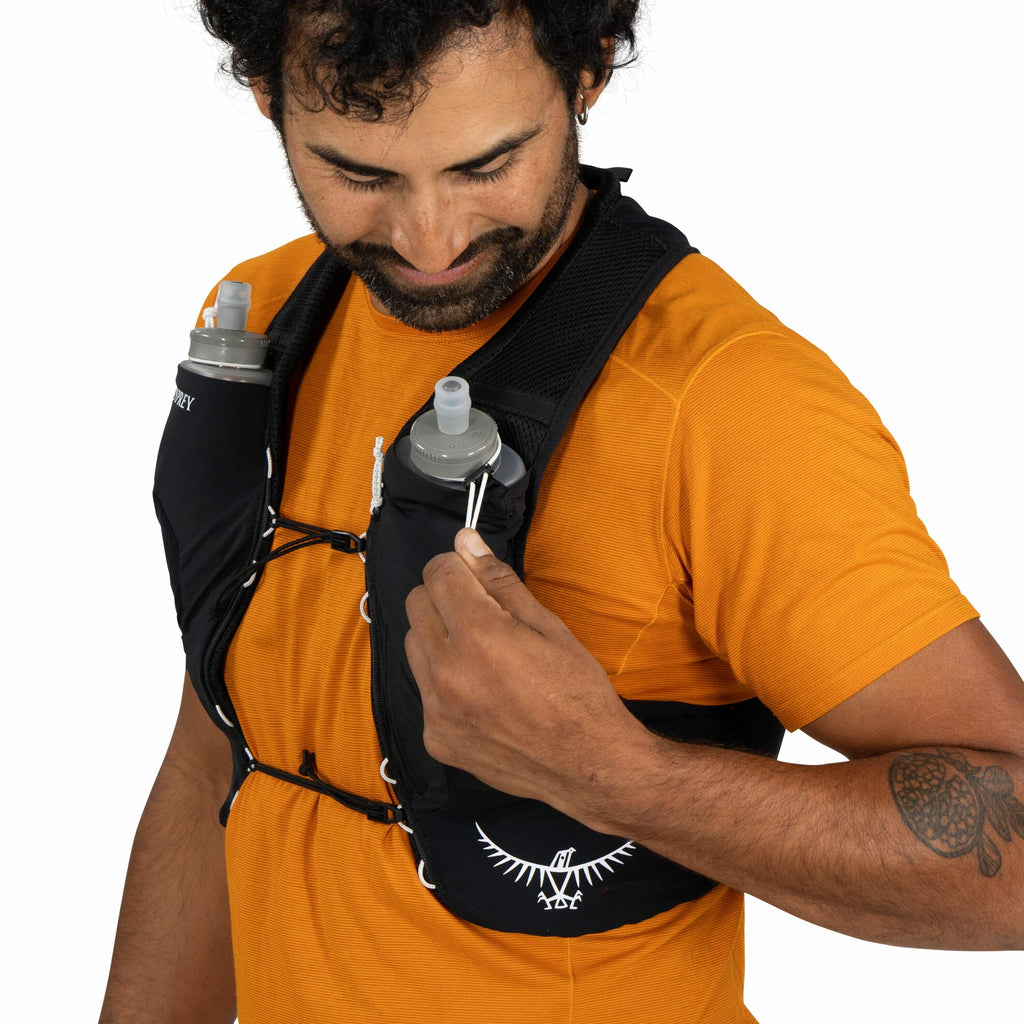 The image shows a person wearing a black hydration vest with two water bottles inserted in front pockets. The vest features the Osprey logo on one side, which is distinctive with its bird silhouette and outstretched wings. The person is dressed in an oran