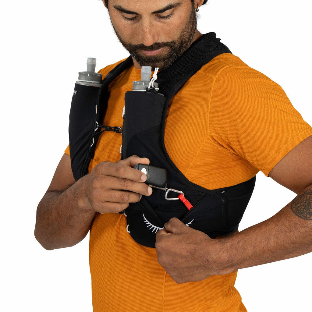 The image shows a person wearing a black running vest with two water bottles attached to the front. The vest is securely fastened with adjustable straps and features a few other pockets for storage. The person is wearing an orange short-sleeved athletic s