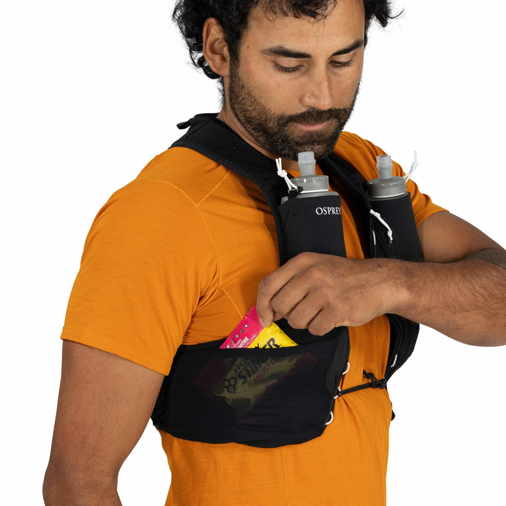 The image shows a person wearing a bright orange t-shirt and a black hydration vest. The vest is equipped with two grey water bottles, each having a white spout, nestled in compartments on the front. The person is reaching into a mesh pocket on the vest's