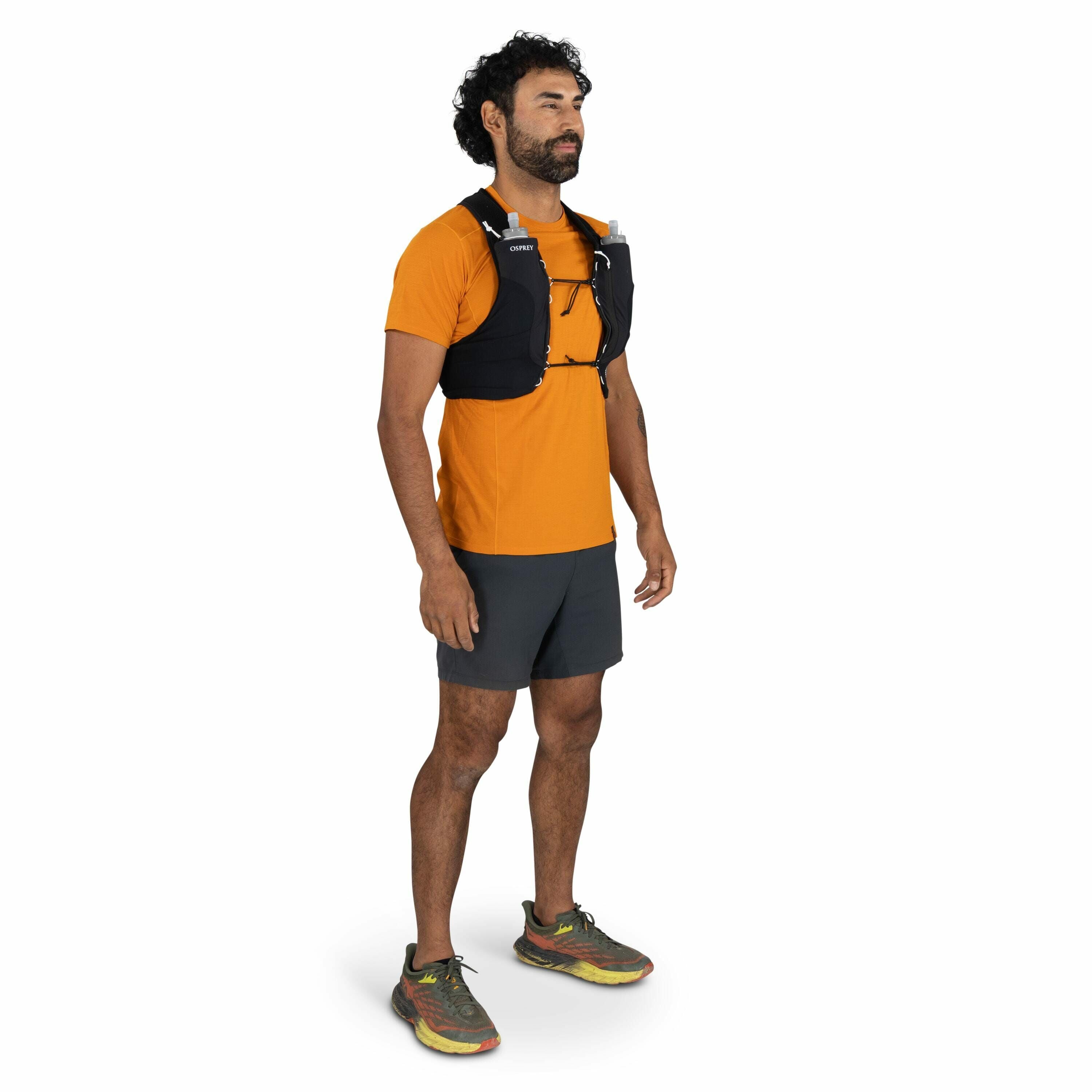 The image features a person standing in a casual and relaxed posture. They are wearing an orange short-sleeve athletic shirt and dark gray shorts, suitable for outdoor activities such as running or hiking. On the person's torso, there is a black hydration