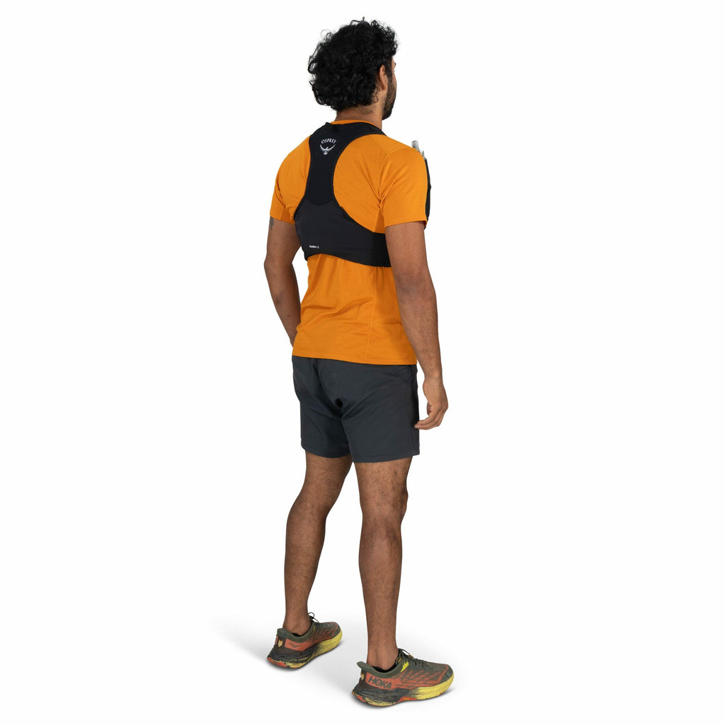 The image shows a man standing in profile, slightly angled away from the camera, wearing an orange short-sleeve shirt and dark gray athletic shorts. On his back, he has a black hydration vest from the brand Osprey, with the logo visible near the upper bac