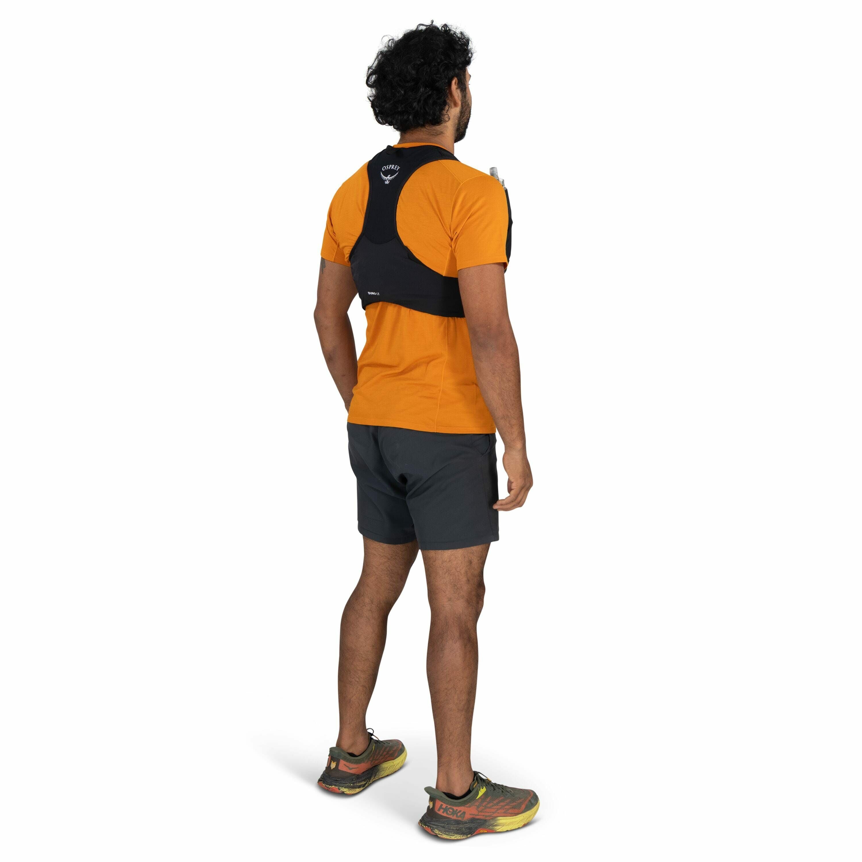 The image shows a man standing in profile, slightly angled away from the camera, wearing an orange short-sleeve shirt and dark gray athletic shorts. On his back, he has a black hydration vest from the brand Osprey, with the logo visible near the upper bac
