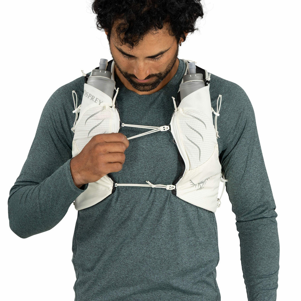 The image shows a person wearing a light grey hydration vest over a dark teal, long-sleeved shirt. The vest is equipped with two water bottles, one on each side of the chest area. The bottles are marked with the "OSPREY" logo, indicating they are likely f