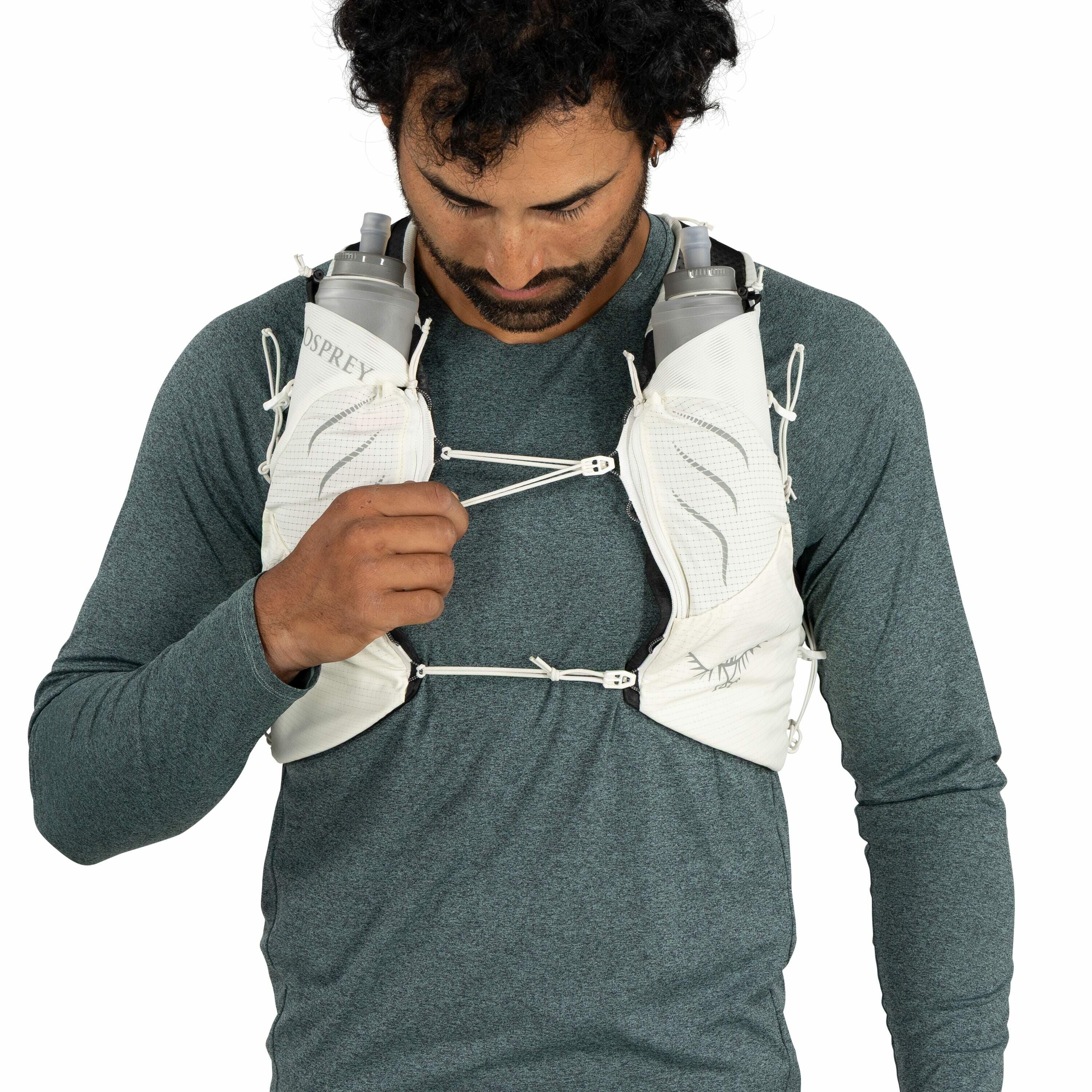 The image shows a person wearing a light grey hydration vest over a dark teal, long-sleeved shirt. The vest is equipped with two water bottles, one on each side of the chest area. The bottles are marked with the "OSPREY" logo, indicating they are likely f