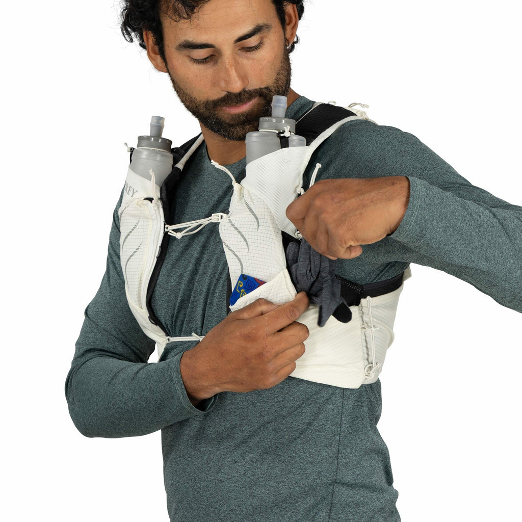 The image shows a person dressed in a long-sleeved green top, wearing a white running vest, designed for hydration and carrying essential items during activities like trail running. The vest has two large front pockets that are equipped with water bottles