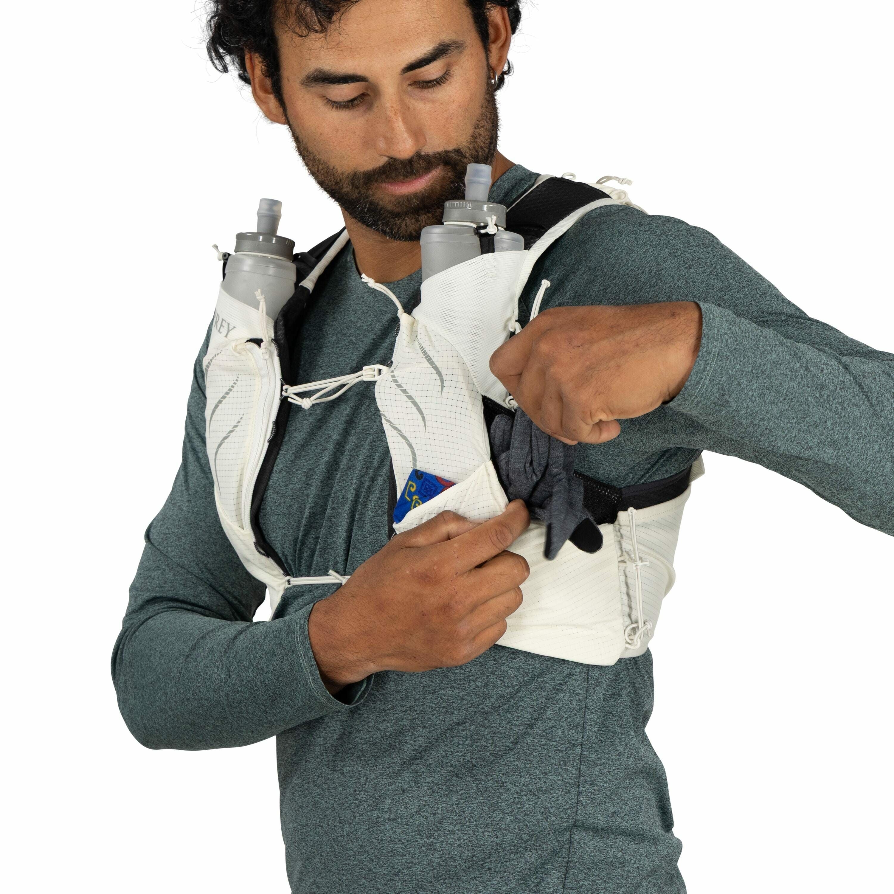 The image shows a person dressed in a long-sleeved green top, wearing a white running vest, designed for hydration and carrying essential items during activities like trail running. The vest has two large front pockets that are equipped with water bottles