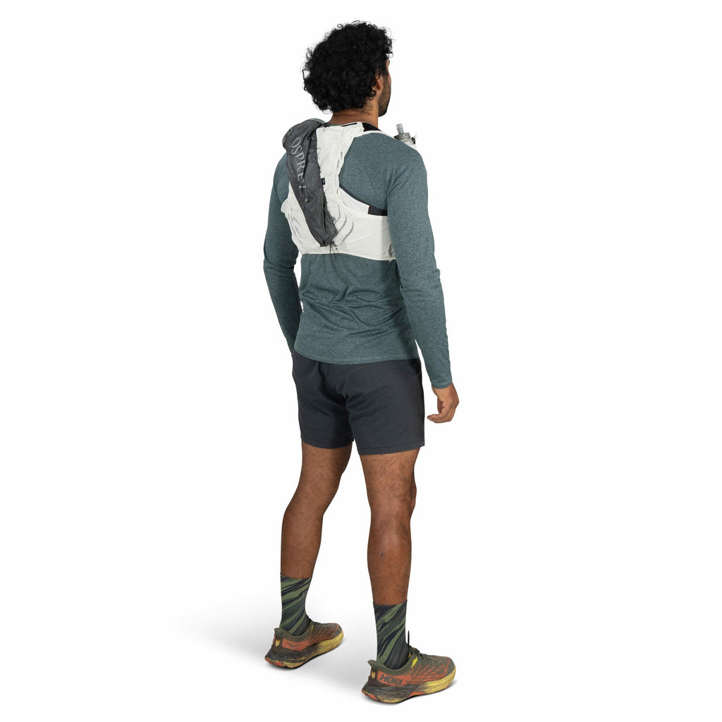 The image shows a man standing and facing away from the camera, wearing a green long-sleeve shirt paired with dark shorts. He has curly black hair and is sporting green and black patterned socks with a pair of brown and orange Hoka running shoes. On his b