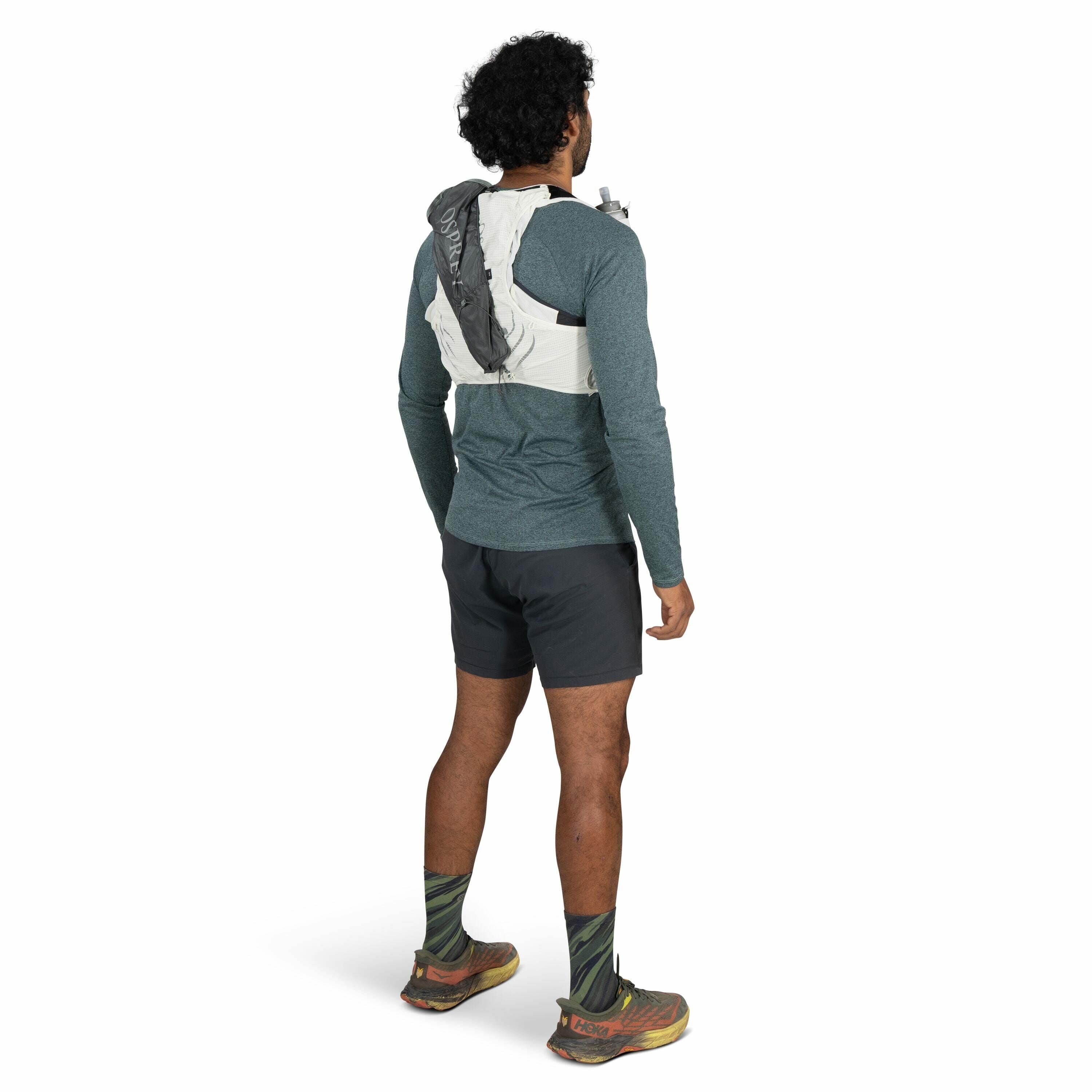 The image shows a man standing and facing away from the camera, wearing a green long-sleeve shirt paired with dark shorts. He has curly black hair and is sporting green and black patterned socks with a pair of brown and orange Hoka running shoes. On his b