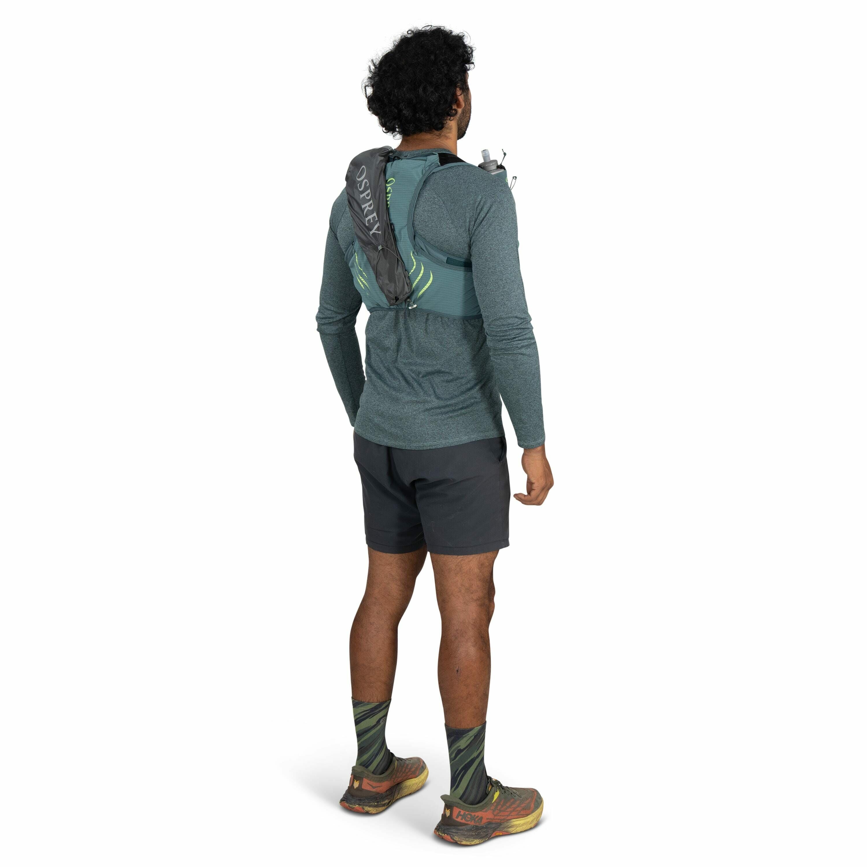 The image shows a man standing with his back to the camera, dressed in outdoor athletic gear. He is wearing a long-sleeved teal shirt, dark gray shorts, and green patterned socks with trail running shoes that have the HOKA logo visible on the back. Over h
