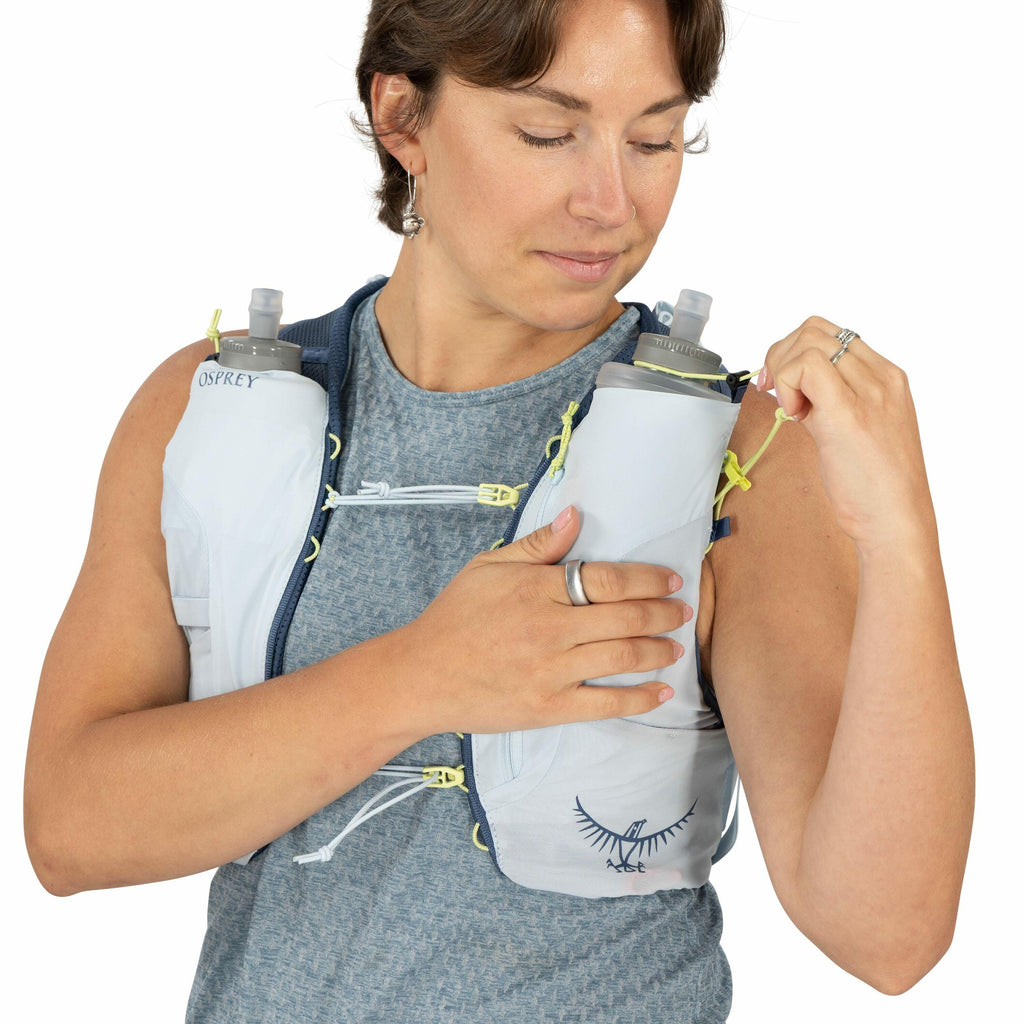 The image depicts a person wearing an Osprey hydration vest, which is designed for activities like running or hiking. The vest is light gray with neon yellow accents and has two soft water bottles positioned in pockets on the chest for easy access to hydr