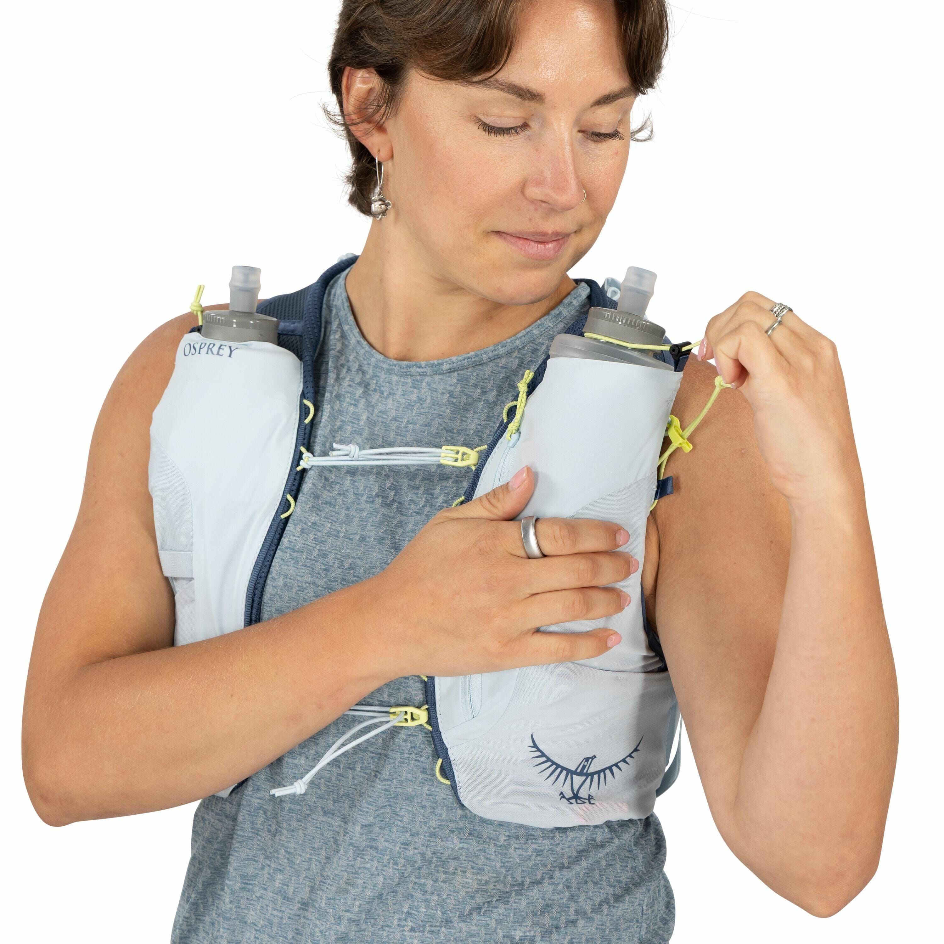 The image depicts a person wearing an Osprey hydration vest, which is designed for activities like running or hiking. The vest is light gray with neon yellow accents and has two soft water bottles positioned in pockets on the chest for easy access to hydr