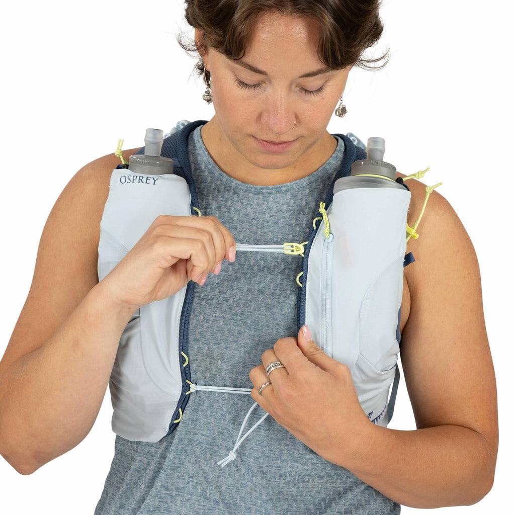 The image depicts a person wearing a light-colored hydration vest by Osprey. The vest is equipped with two water bottles positioned on the front, each secured in a pocket for easy access. The person is holding a strap, which seems to be part of the vest's