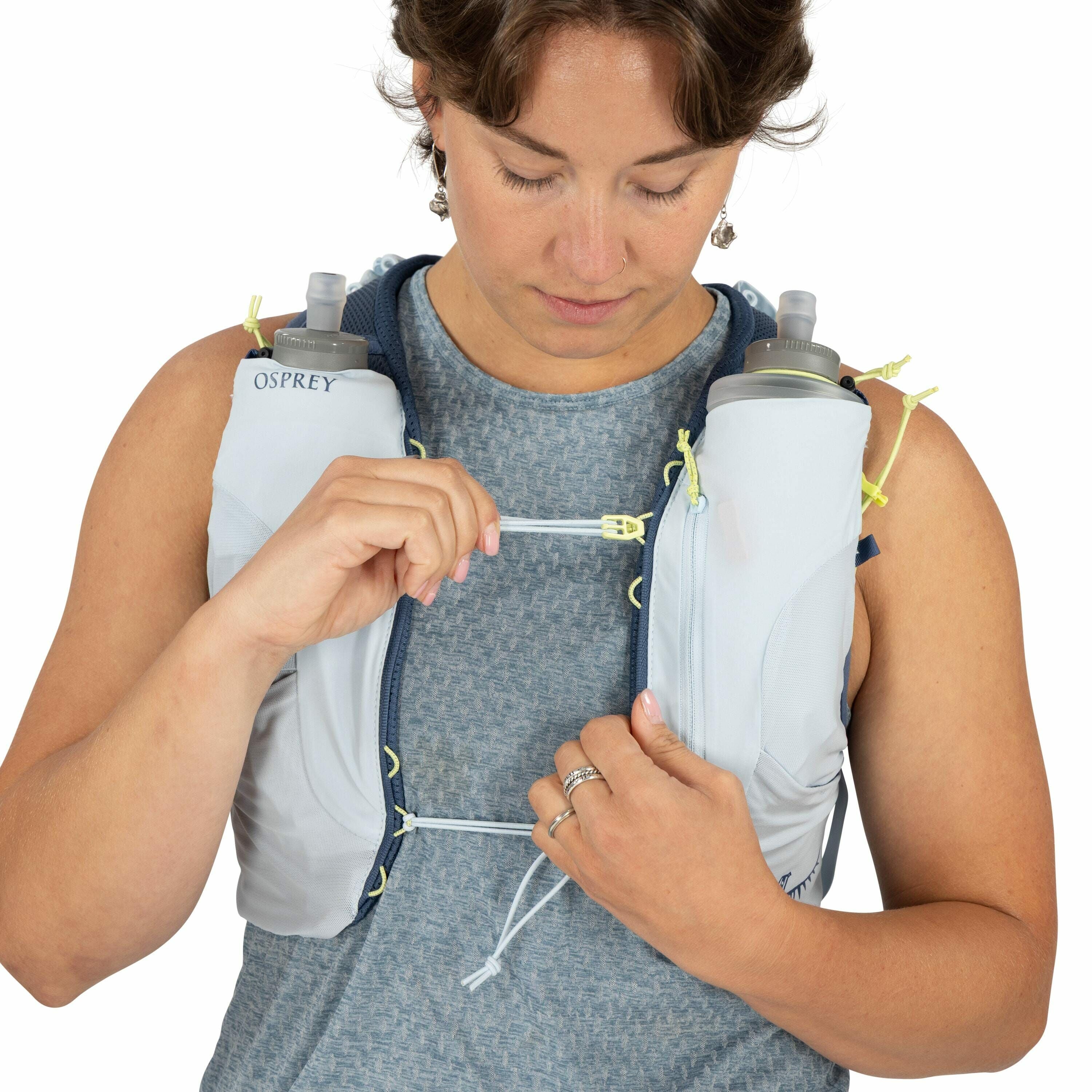 The image depicts a person wearing a light-colored hydration vest by Osprey. The vest is equipped with two water bottles positioned on the front, each secured in a pocket for easy access. The person is holding a strap, which seems to be part of the vest's