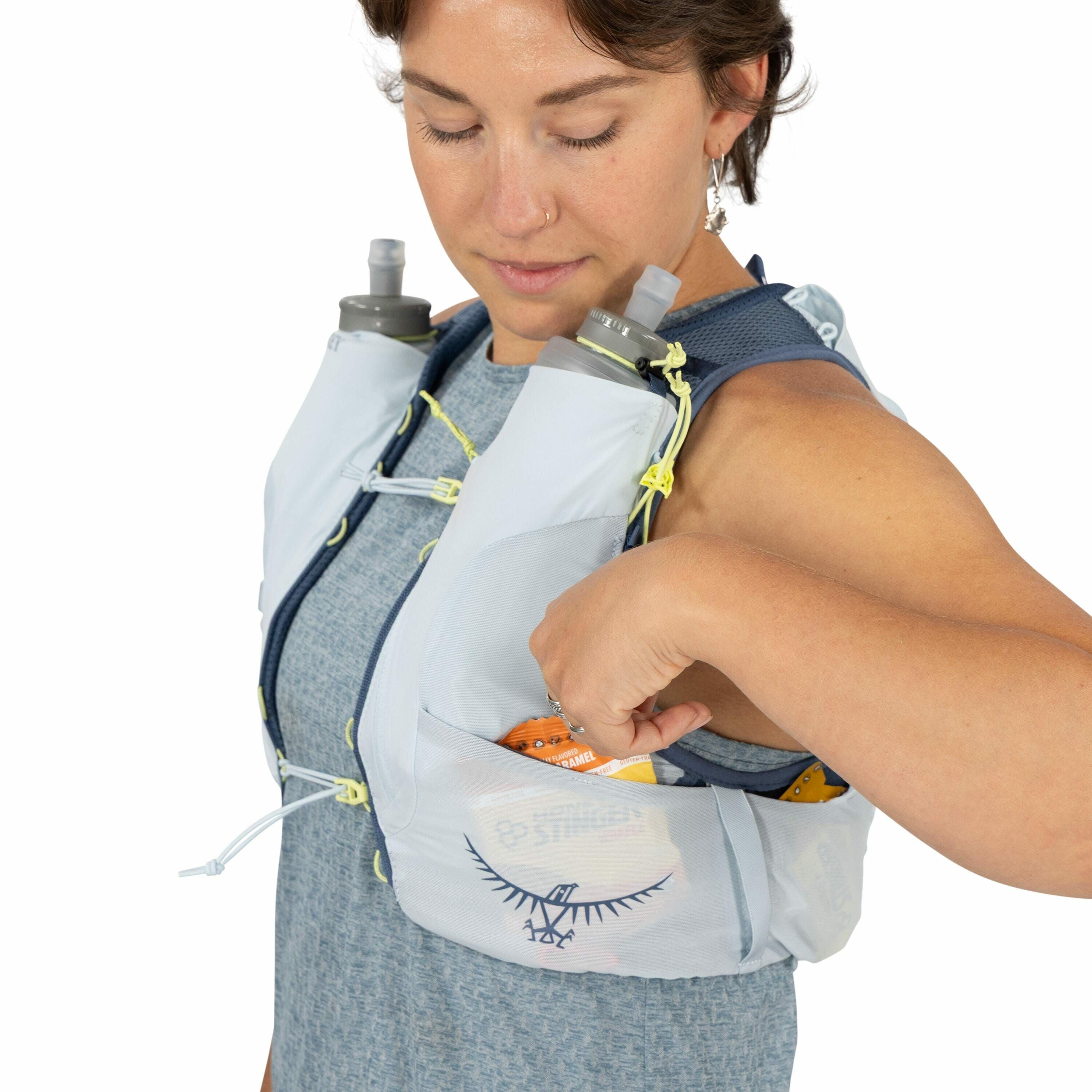 The image shows a person wearing a light-colored hydration vest designed for outdoor activities, such as running or hiking. The vest is equipped with two water bottles secured in front pockets. There are visible strings and adjustable straps on the vest f