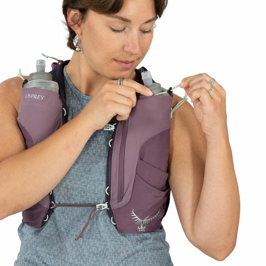 The image shows a person wearing a purple Osprey hydration vest over a grey textured sleeveless shirt. The vest features two visible soft water bottles, which are stored in mesh pockets on the chest area of the vest. The individual is adjusting a drawstri