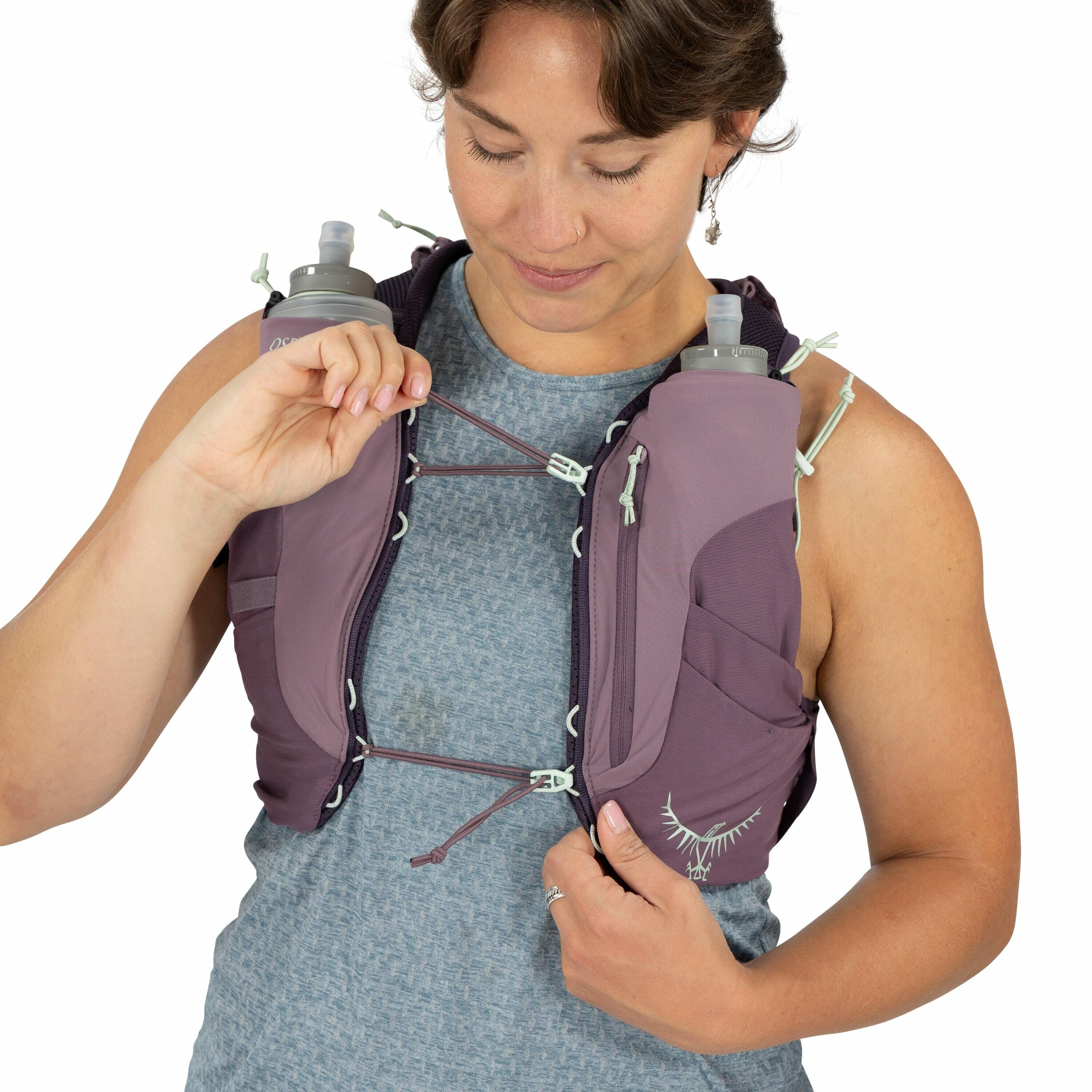 The image features a person wearing a hydration vest, which is prominently displayed on their upper torso. The vest is purple with light green accents and is equipped with two water bottles, one on each side of the chest. The person is adjusting the vest