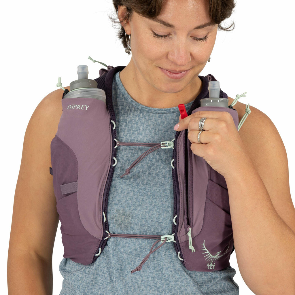 The image shows a person wearing a purple Osprey running hydration vest. The vest has adjustable cords across the chest, securing the fit. It contains two soft flasks, one on each side of the vest, which are accessible for drinking while running. The pers