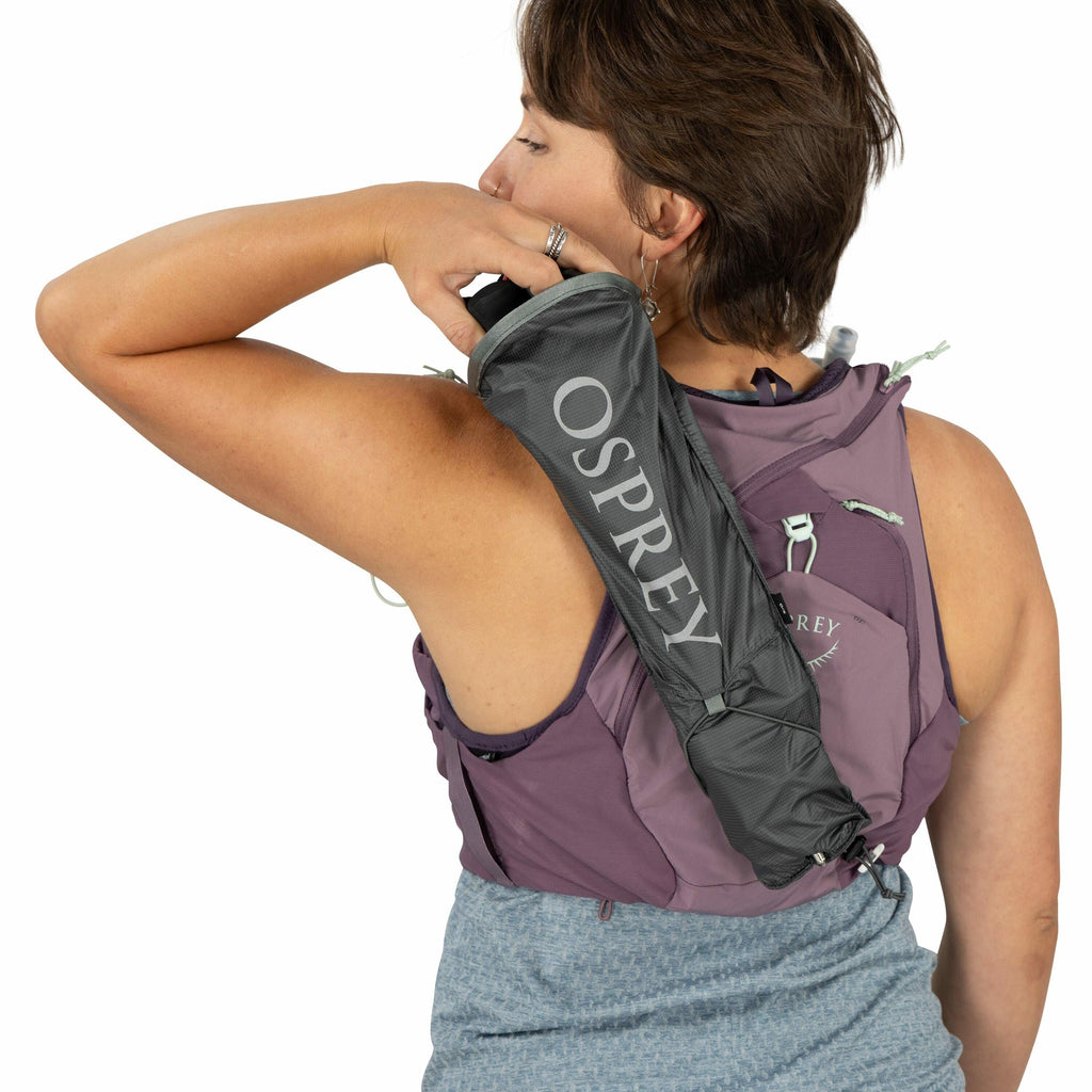 The image shows a person from behind, wearing a purple backpack that appears to be from the Osprey brand, as indicated by the logo. The person is also holding an Osprey rain cover or jacket over their shoulder. The backpack features multiple pockets and s