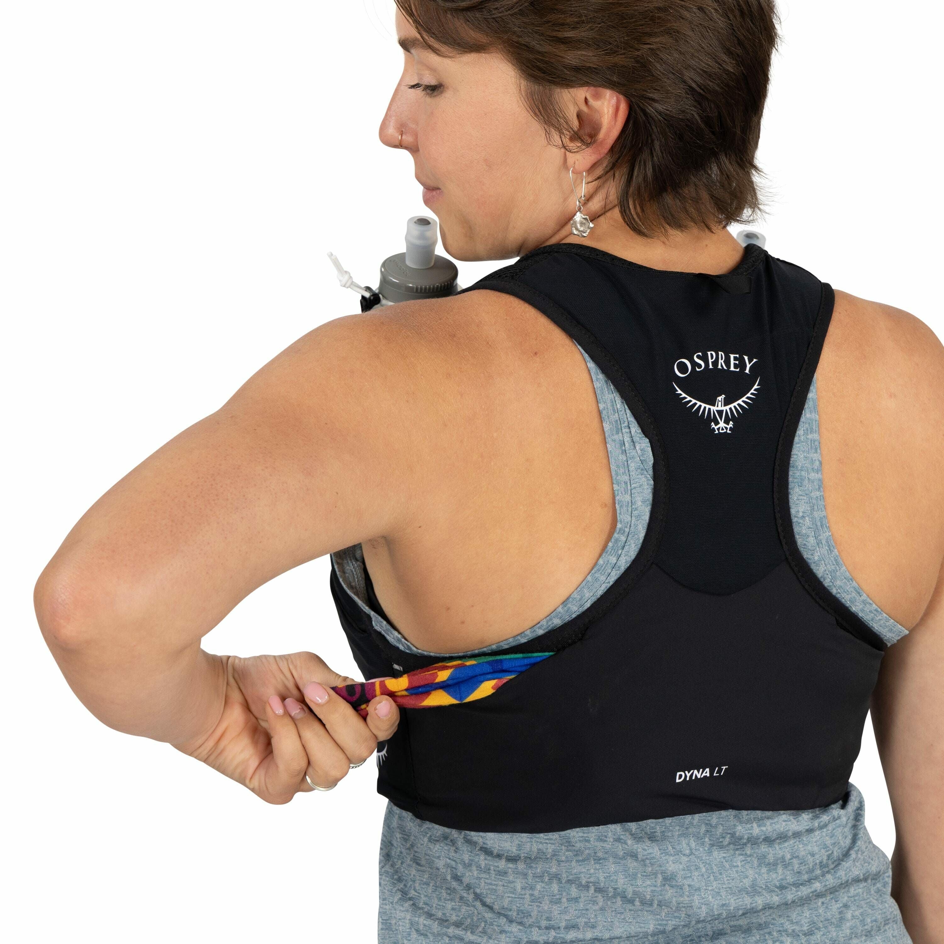 The image features a person wearing an Osprey brand hydration vest with the logo visible on the back. The vest, labeled as "DYNA LT," is black and fitted closely to the personÕs body, over a grey-blue patterned shirt. The person is using one hand to place