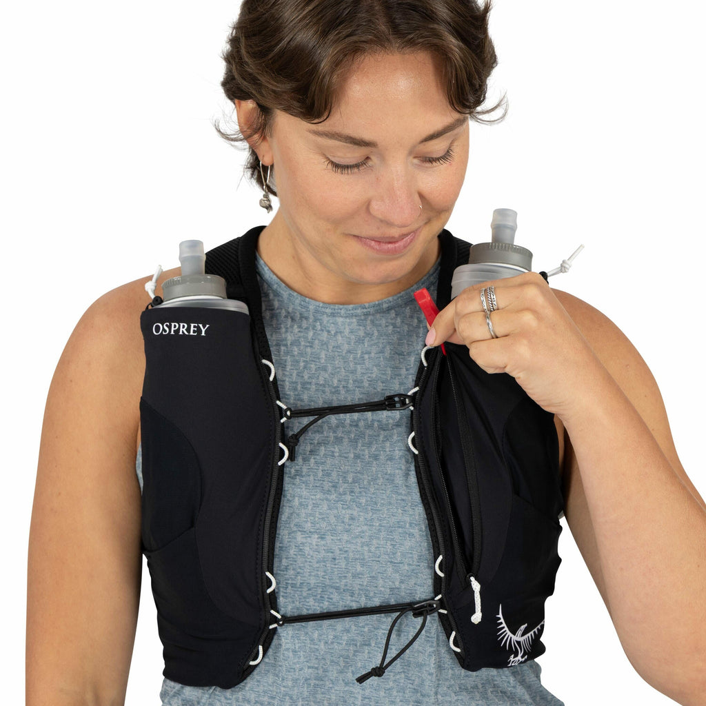 The image shows a person wearing a black hydration vest from the brand Osprey. The vest is equipped with two soft water bottles, each inserted into pockets on the front of the vest. The person is wearing a sleeveless, textured blue-gray top underneath the