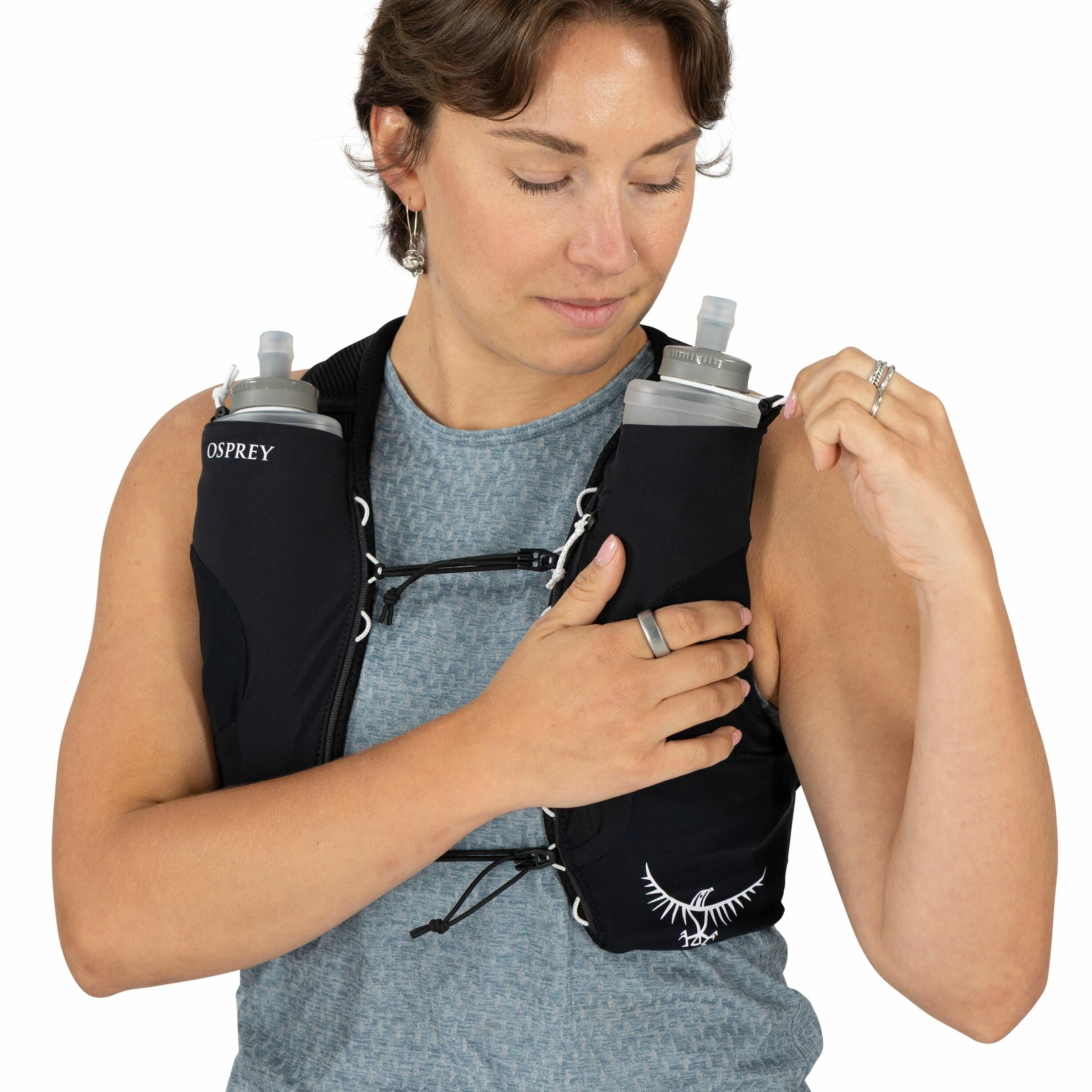 The image shows a person wearing an Osprey hydration vest, which is designed for easy access to fluids during activities like running or hiking. The vest is black and features two built-in water bottle holders on the front, each holding a soft flask with