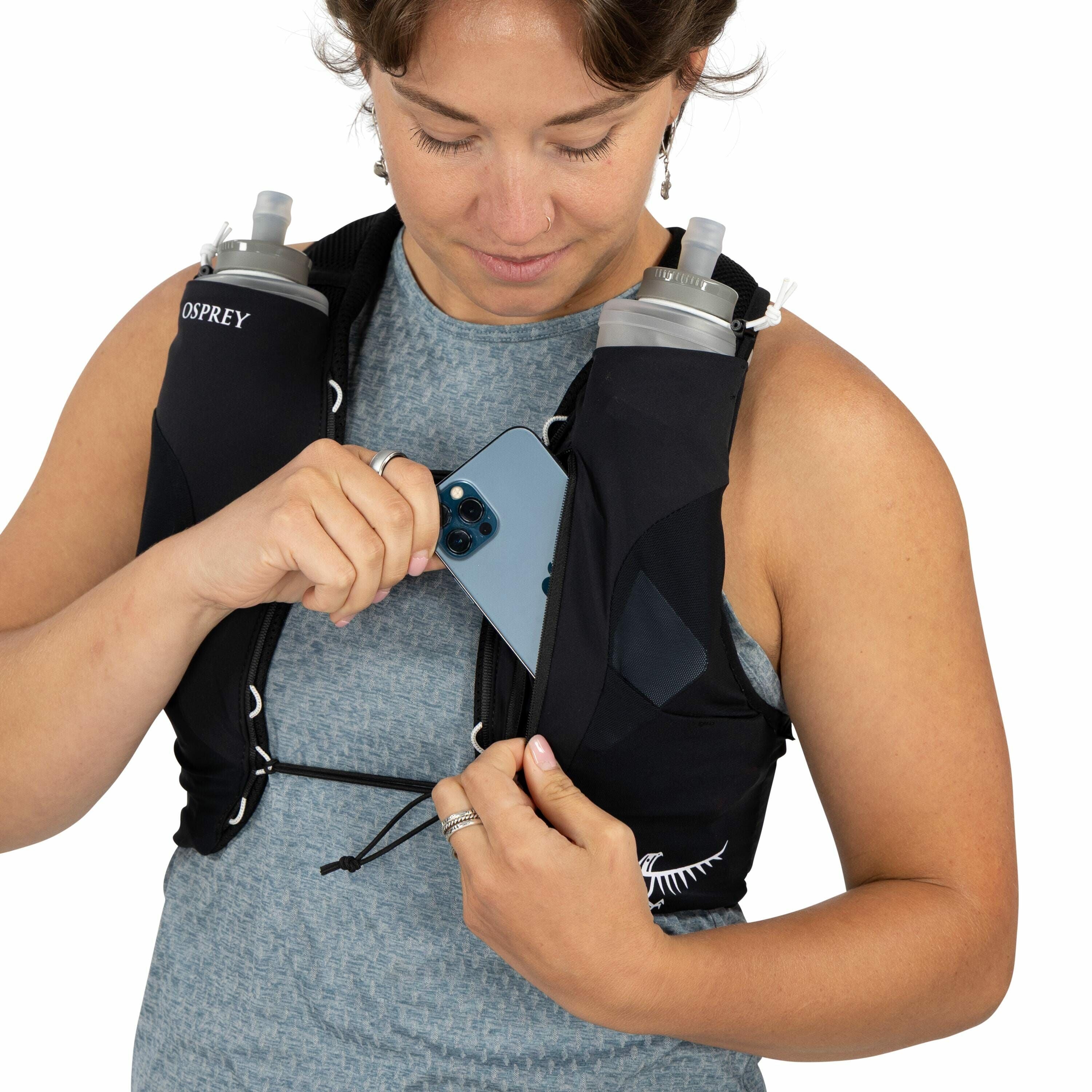 The image shows a person wearing a black hydration vest with two water bottles placed in front pockets on either side of the vest. The vest has the brand name "Osprey" visible near the shoulder area. The individual is holding a smartphone, which appears t