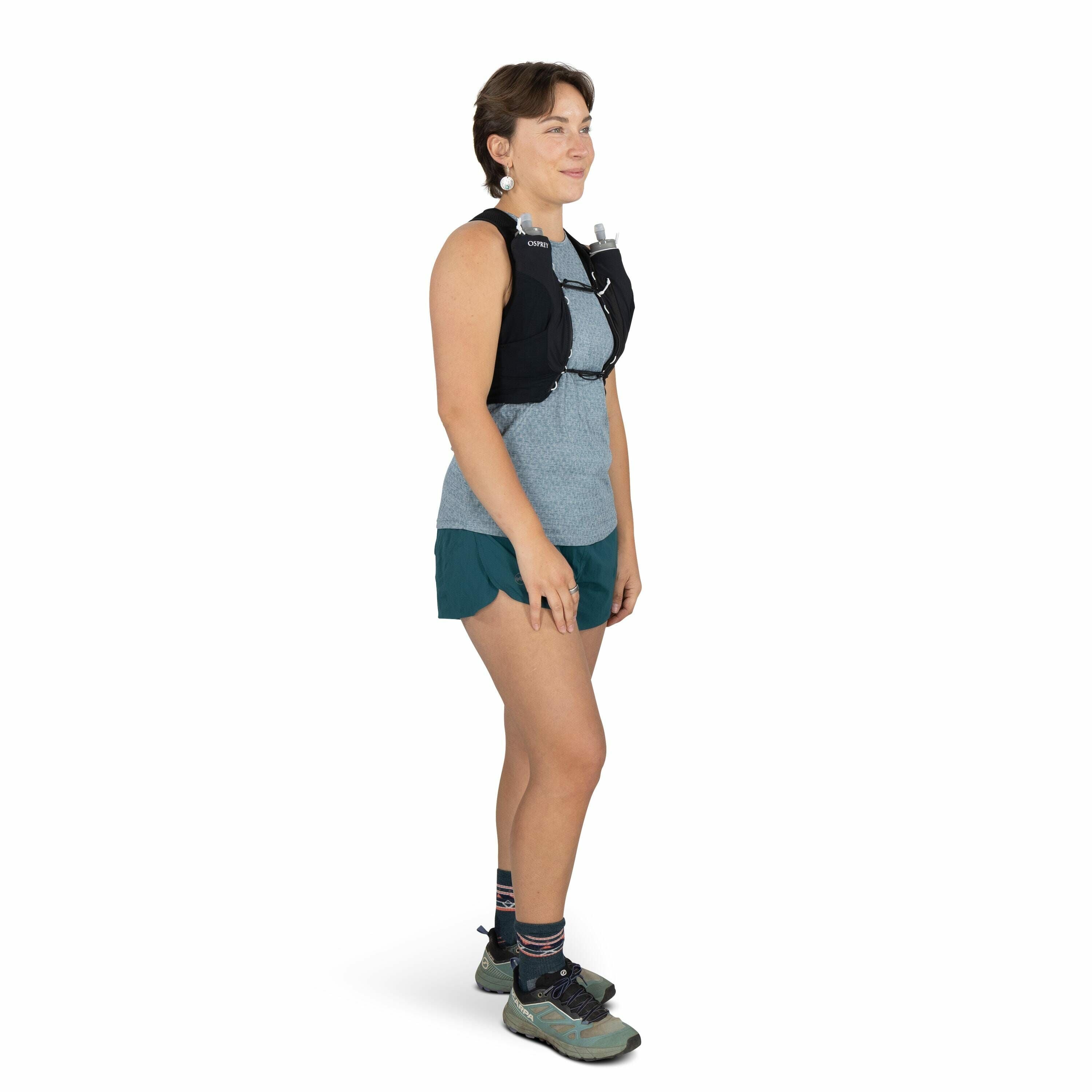 The image shows a person standing sideways to the camera, dressed in outdoor athletic attire suitable for activities like running or hiking. They are wearing a blue-gray patterned sleeveless top and dark teal athletic shorts. The person has a black hydrat