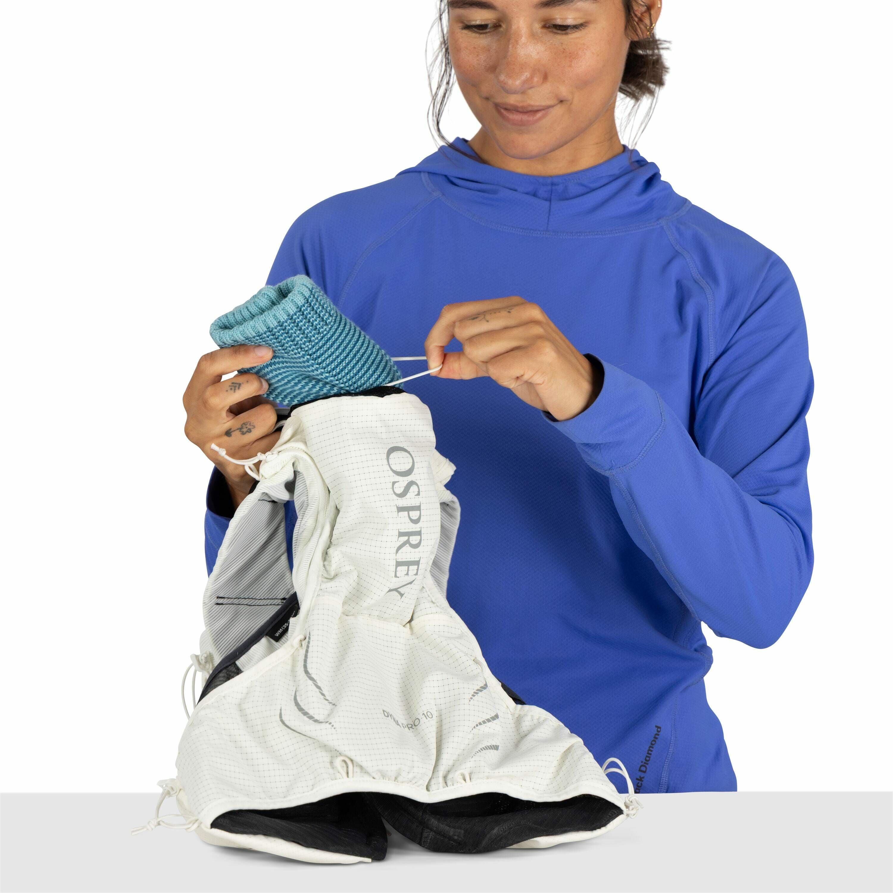 The image shows a person wearing a vibrant blue long-sleeve top with a hood. They are holding an Osprey backpack, which is primarily white with some black details. The person is in the process of inserting a rolled, teal-colored knit object into the backp