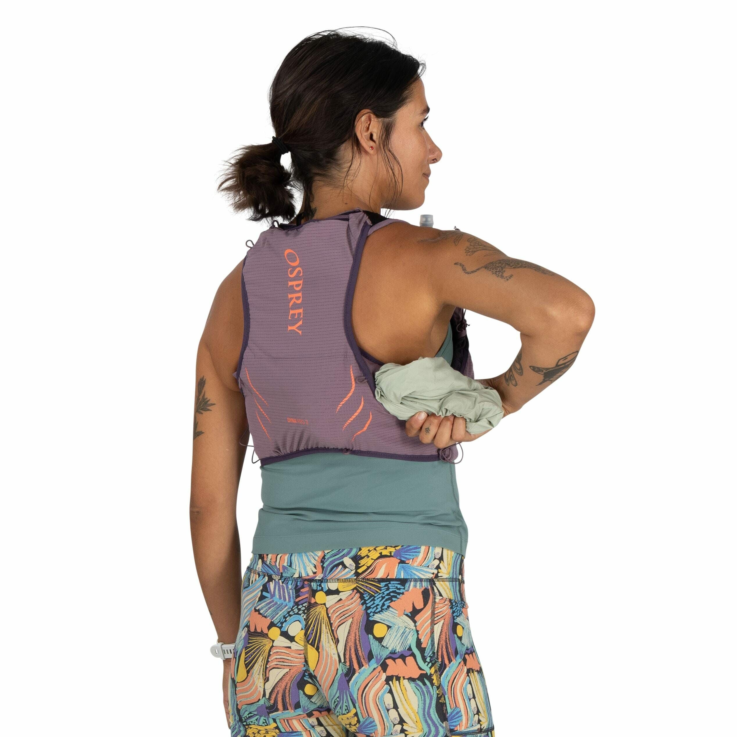 The image shows a person wearing a running vest with the logo "OSPREY" in orange letters on the back. The vest is purple and designed for carrying gear, and the individual is holding a rolled-up piece of clothing in their left hand. The person is dressed