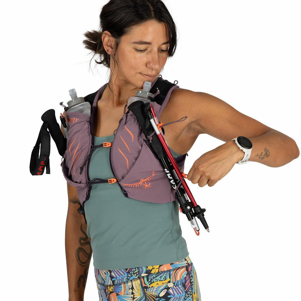 The image shows a person wearing a purple and orange Osprey hydration vest. Attached to the vest are two water bottles, and the person is adjusting a pair of CAMP trekking poles that are secured to the vest's front. They are dressed in a teal tank top and