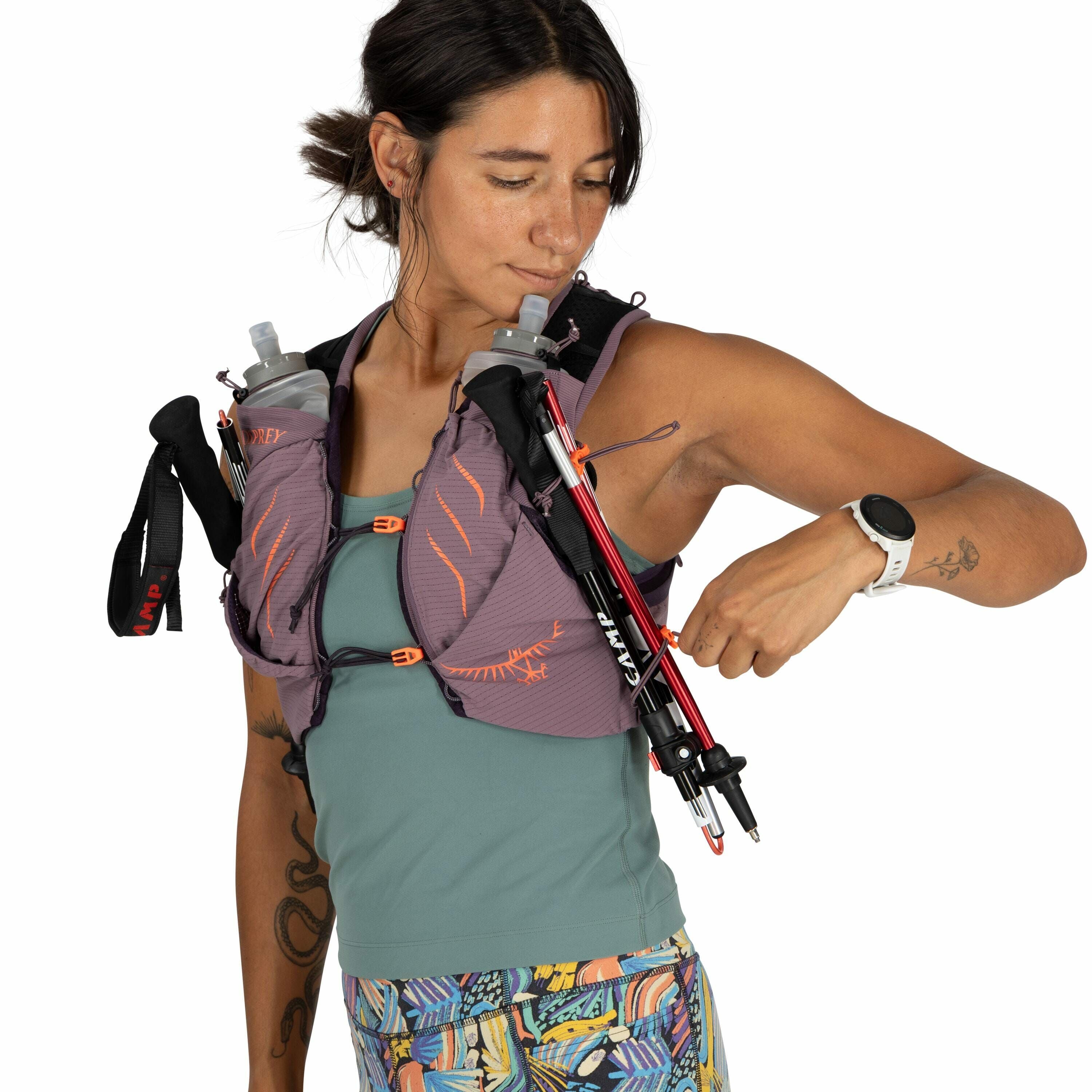 The image shows a person wearing a purple and orange Osprey hydration vest. Attached to the vest are two water bottles, and the person is adjusting a pair of CAMP trekking poles that are secured to the vest's front. They are dressed in a teal tank top and