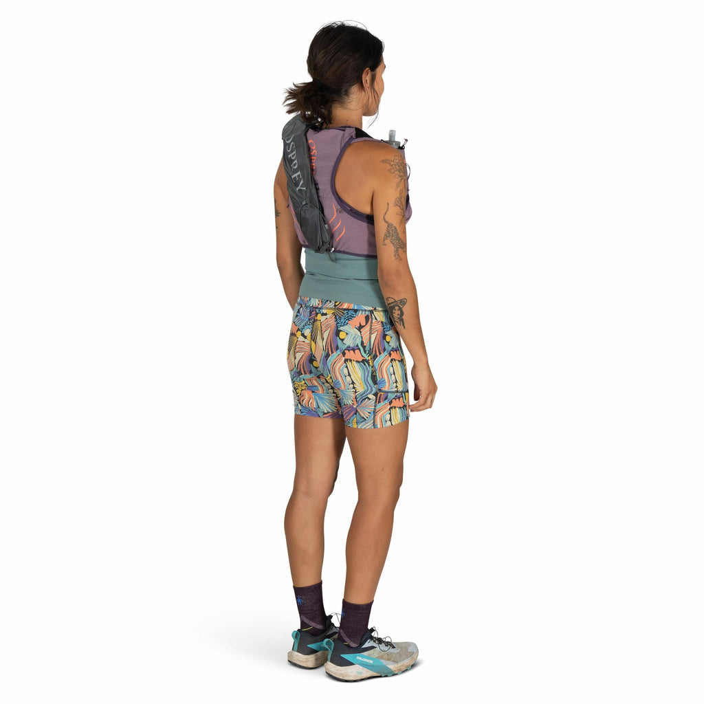 The image shows a person standing and facing away from the camera. They are wearing a sleeveless purple top, colorful shorts with a vibrant pattern featuring abstract shapes, and a pair of running shoes with teal accents. Over their top, they have a hydra