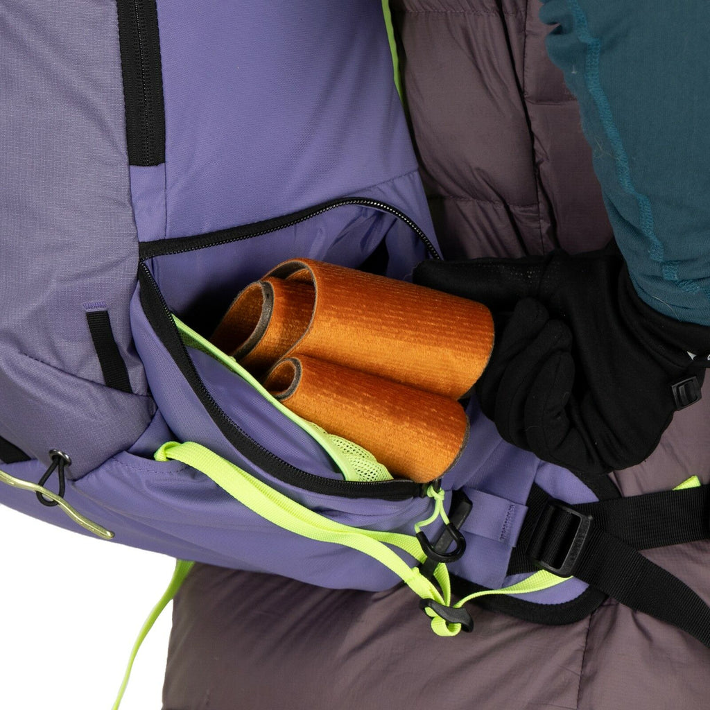 The image shows a close-up of a person carrying a backpack, focusing on a side pocket that is partially open. Inside the pocket, there are rolled-up orange papers or similar objects visible. The backpack itself is predominantly purple with black accents a