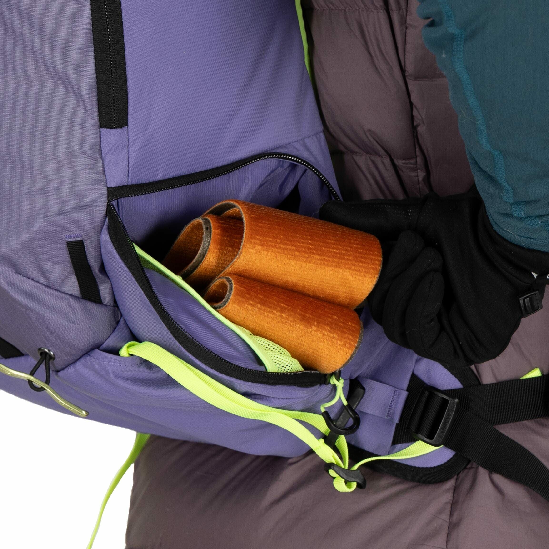 The image shows a close-up of a person carrying a backpack, focusing on a side pocket that is partially open. Inside the pocket, there are rolled-up orange papers or similar objects visible. The backpack itself is predominantly purple with black accents a