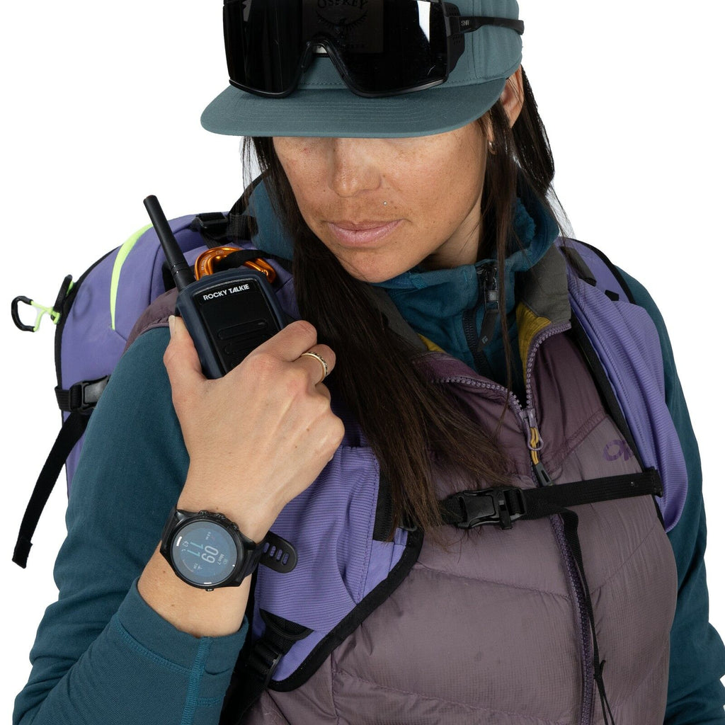 The image shows a person equipped for an outdoor adventure, particularly in a mountainous or snowy environment. They are wearing a purple and teal outdoor jacket with a matching lavender backpack designed for hiking or skiing. On their left wrist, there i