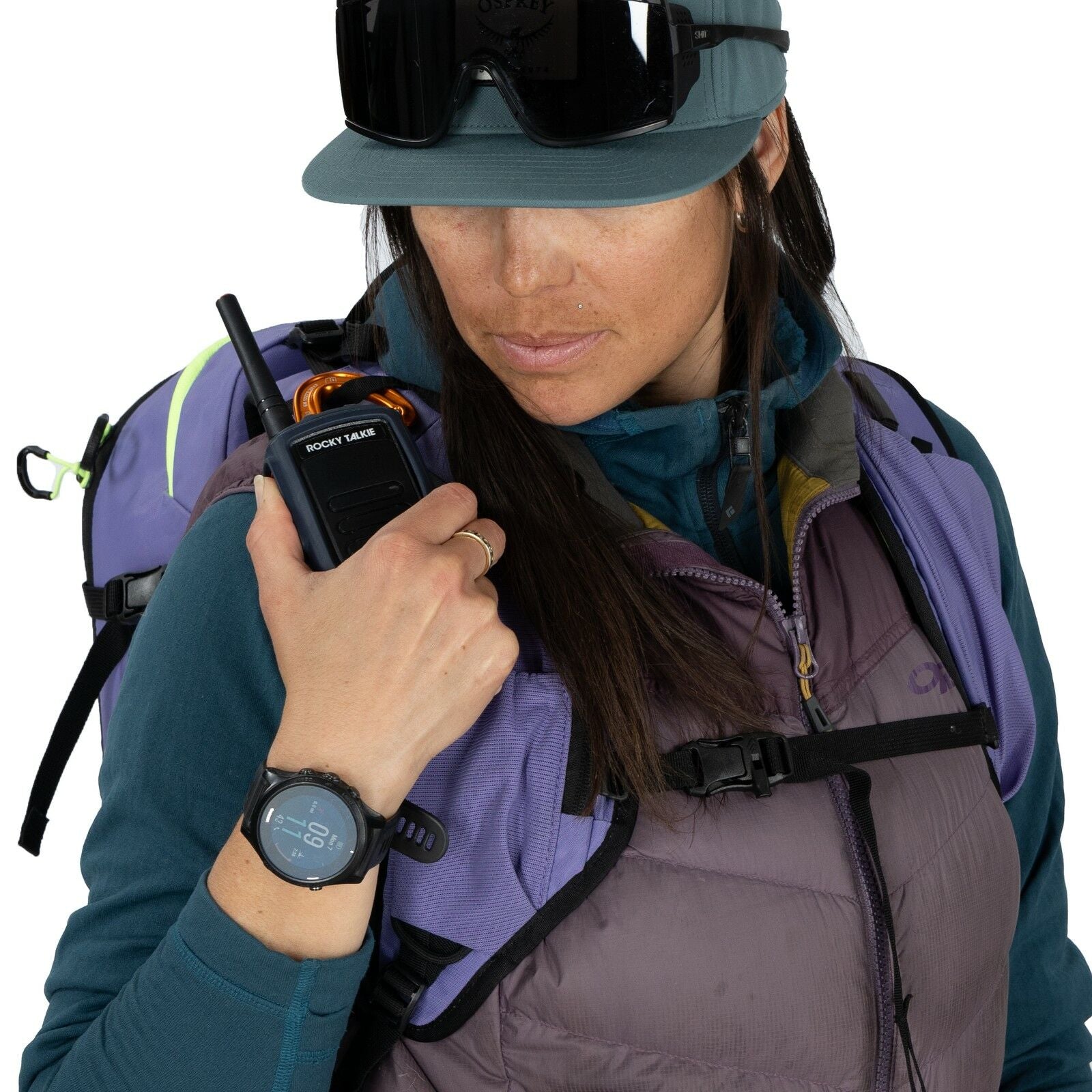 The image shows a person equipped for an outdoor adventure, particularly in a mountainous or snowy environment. They are wearing a purple and teal outdoor jacket with a matching lavender backpack designed for hiking or skiing. On their left wrist, there i