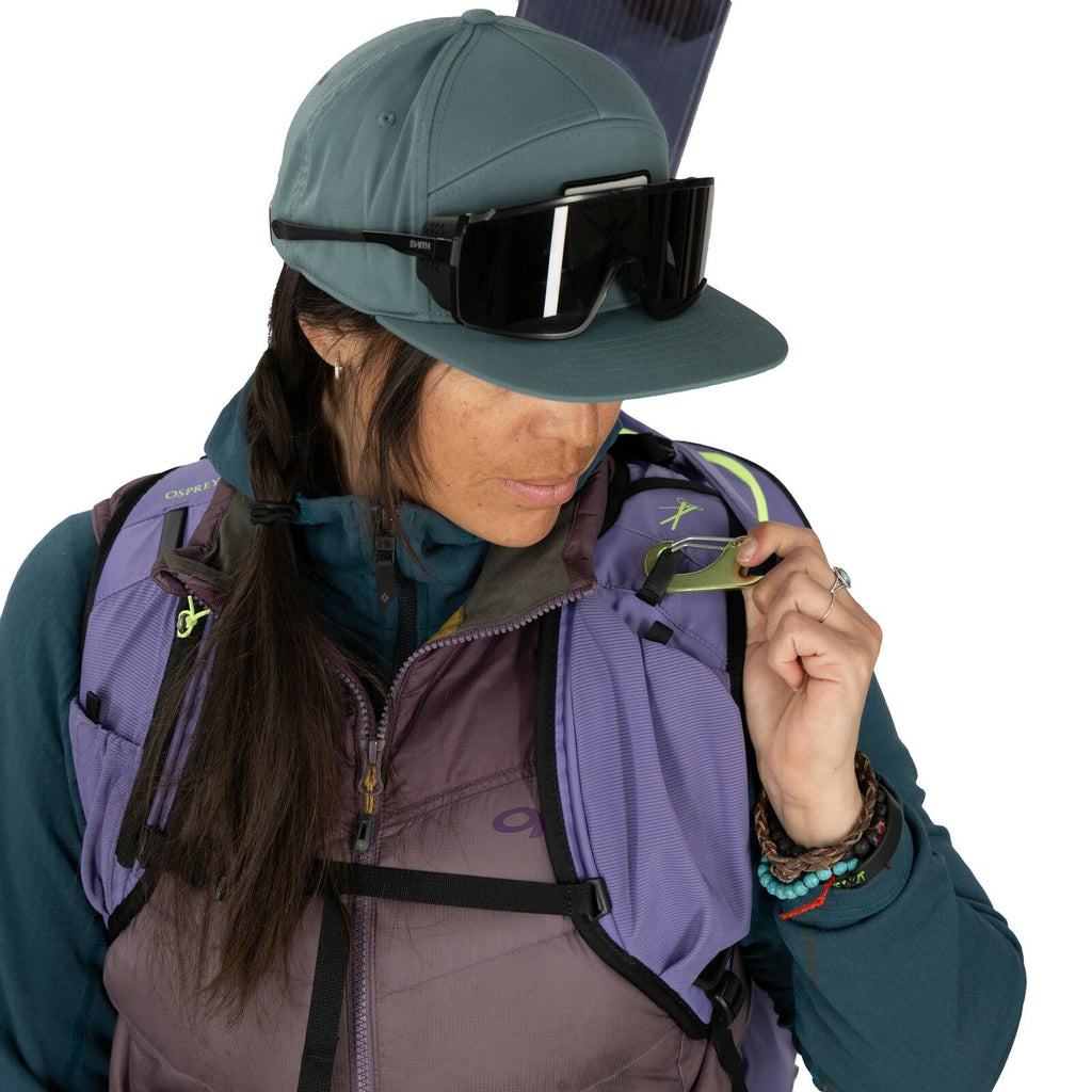 The image shows a person dressed in outdoor and adventure gear, suggesting they are prepared for hiking or a similar activity. They are wearing a blueish-grey cap and large black sunglasses. The individual is also equipped with a purple and grey vest, whi