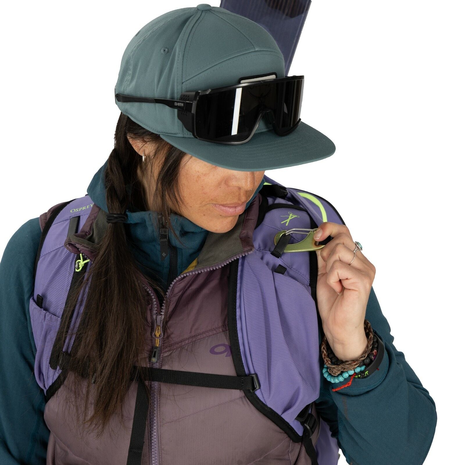 The image shows a person dressed in outdoor and adventure gear, suggesting they are prepared for hiking or a similar activity. They are wearing a blueish-grey cap and large black sunglasses. The individual is also equipped with a purple and grey vest, whi