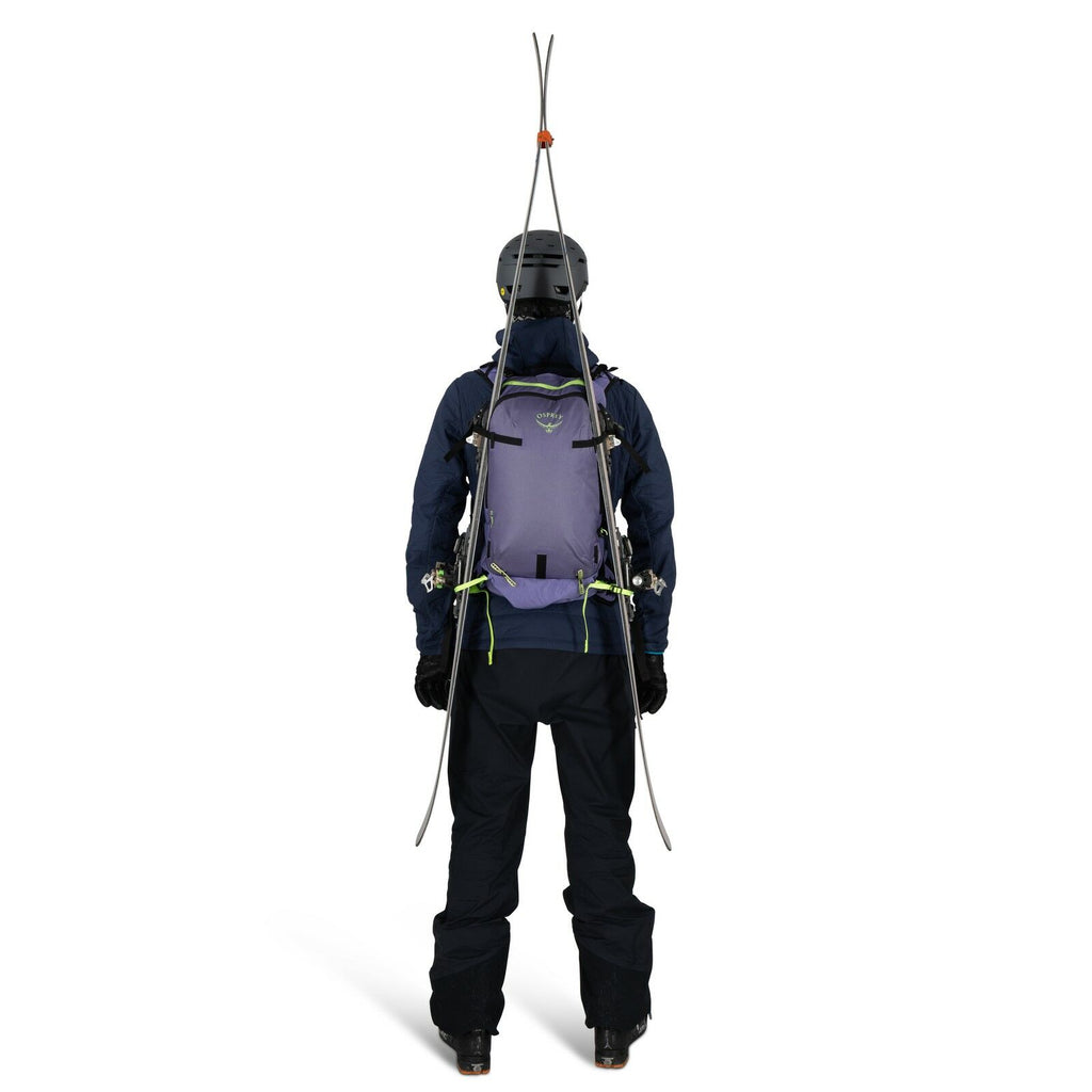 The image shows a person viewed from the back, equipped for skiing or mountaineering. They are wearing a helmet, a dark jacket, and dark pants, with a pair of skis fastened upright to a light purple backpack. The backpack has multiple straps and fastening