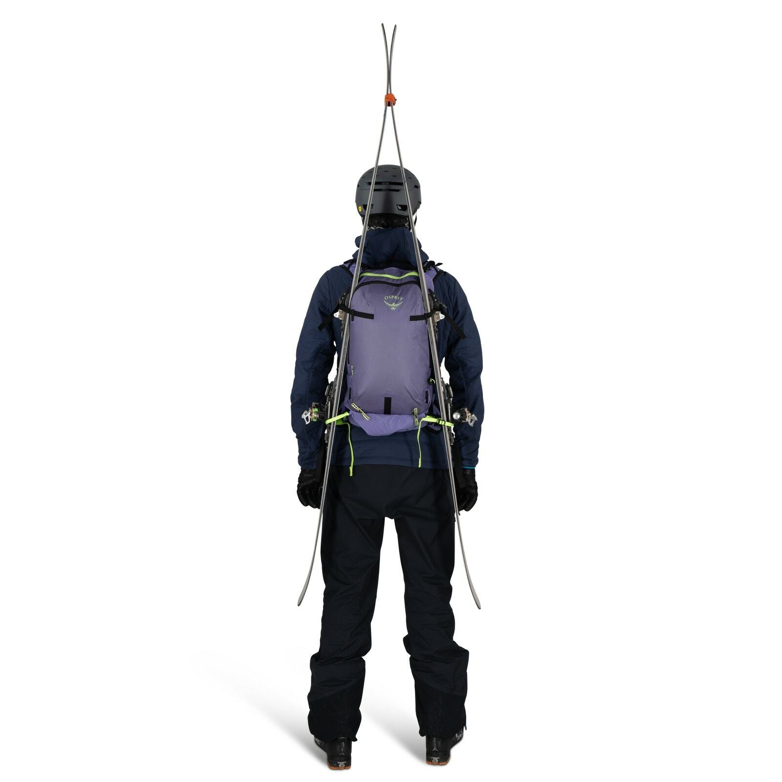 The image shows a person viewed from the back, equipped for skiing or mountaineering. They are wearing a helmet, a dark jacket, and dark pants, with a pair of skis fastened upright to a light purple backpack. The backpack has multiple straps and fastening