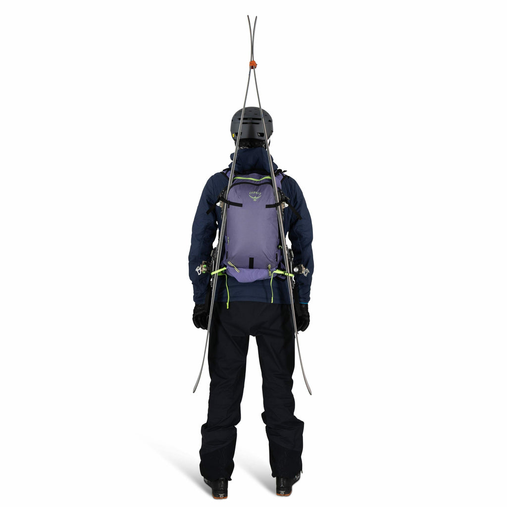 The image shows a person viewed from the back, equipped for skiing or mountaineering. They are wearing a helmet, a dark jacket, and dark pants, with a pair of skis fastened upright to a light purple backpack. The backpack has multiple straps and fastening